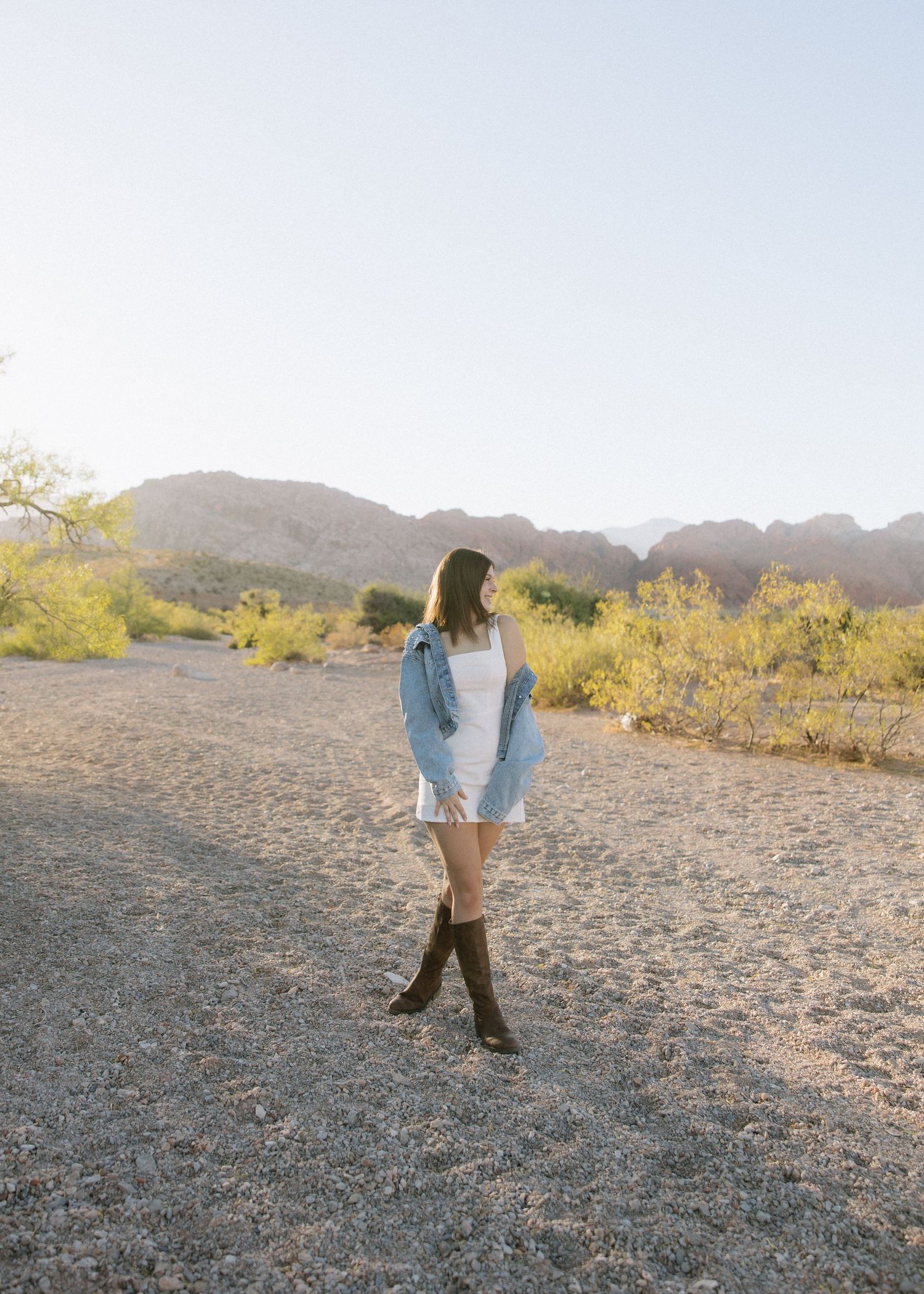 Woman in denim jacket and tall boots poses against desert mountain backdrop.