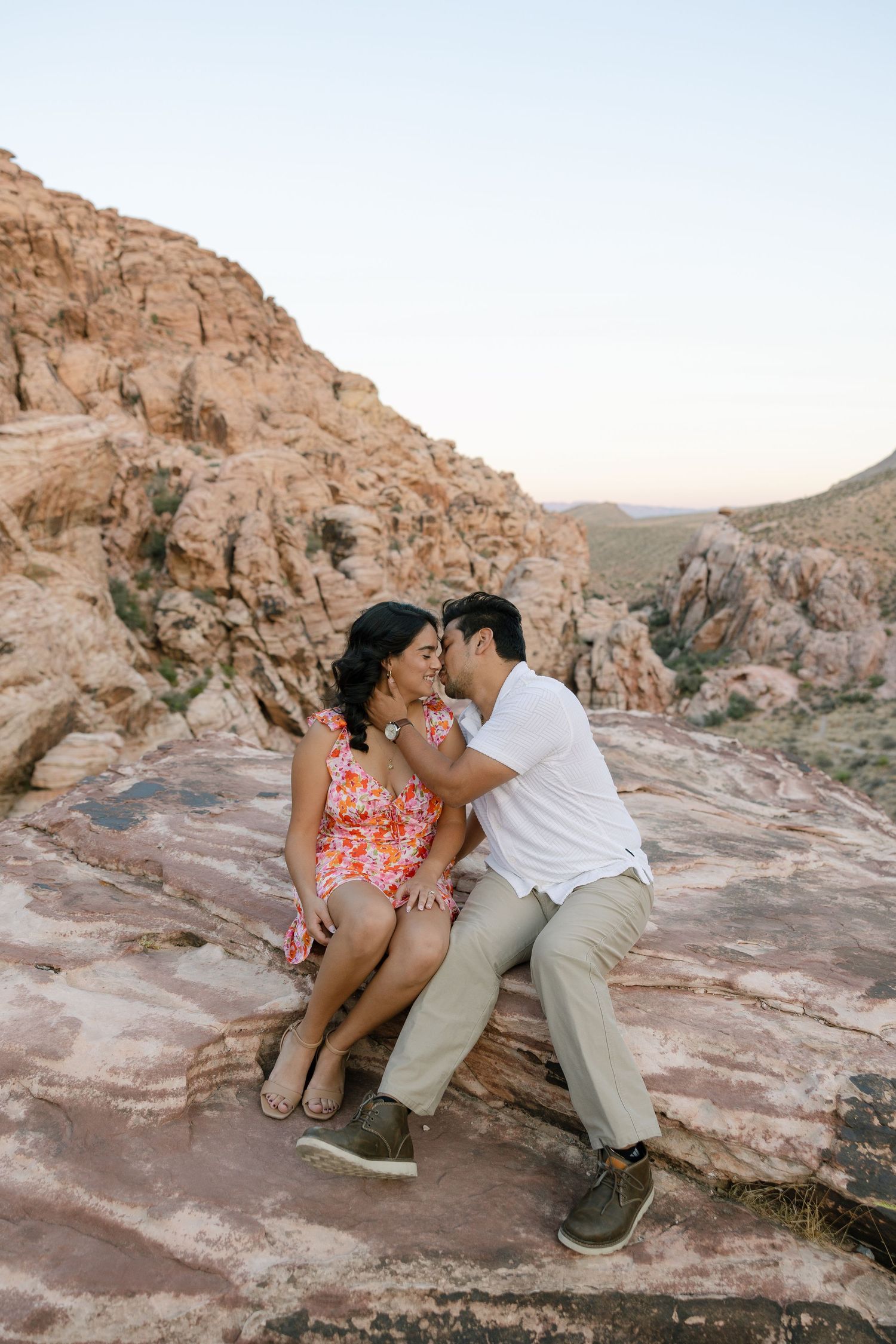 A couple shares an intimate moment while sitting on red rock formations in a desert landscape at sunset.
