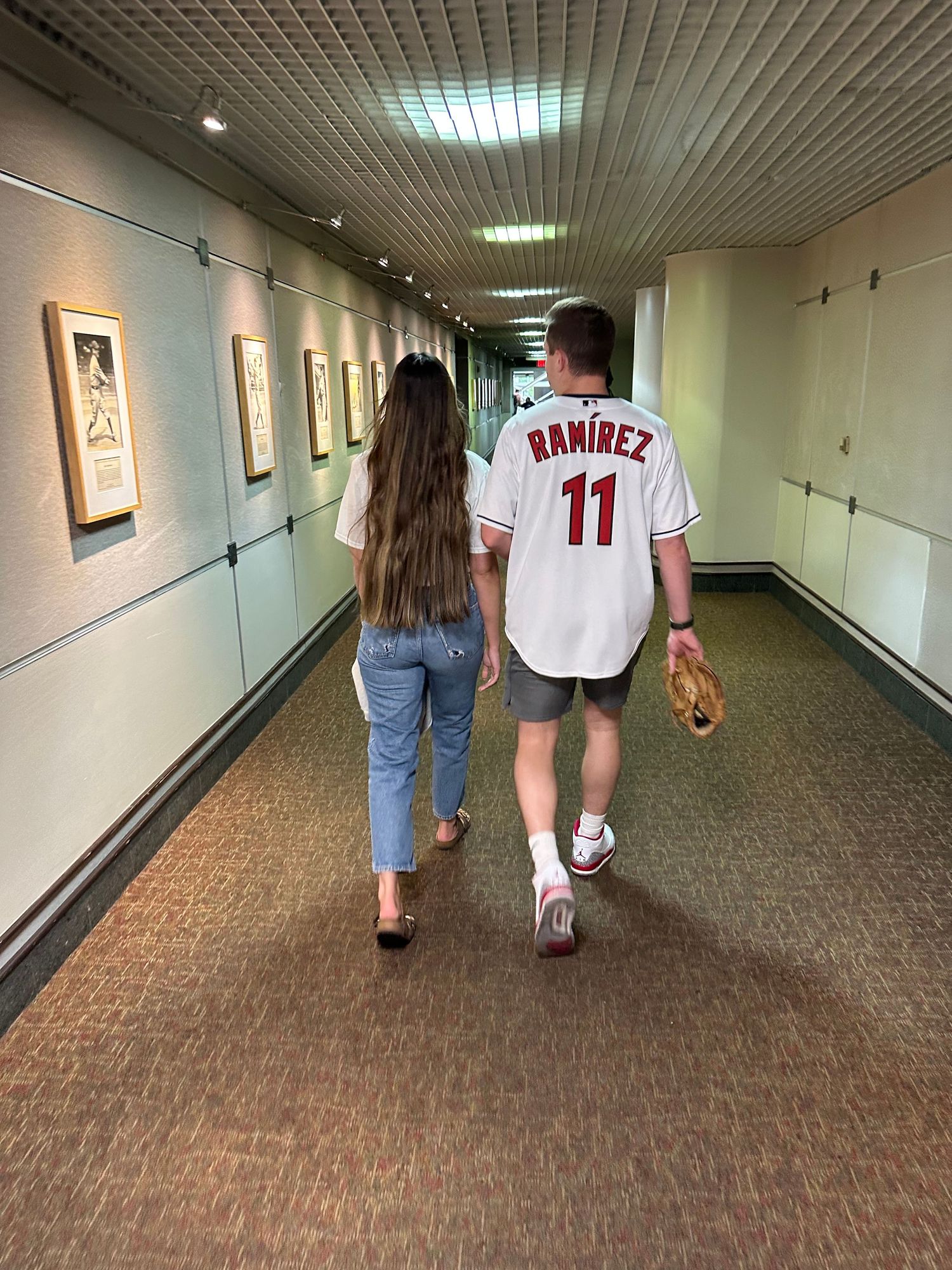 People walking down hallway with baseball jersey and framed artwork on walls.