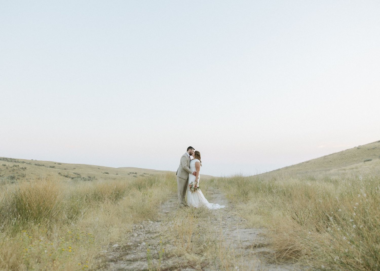 A couple stands together on a winding path through tall grass in an ethereal landscape.