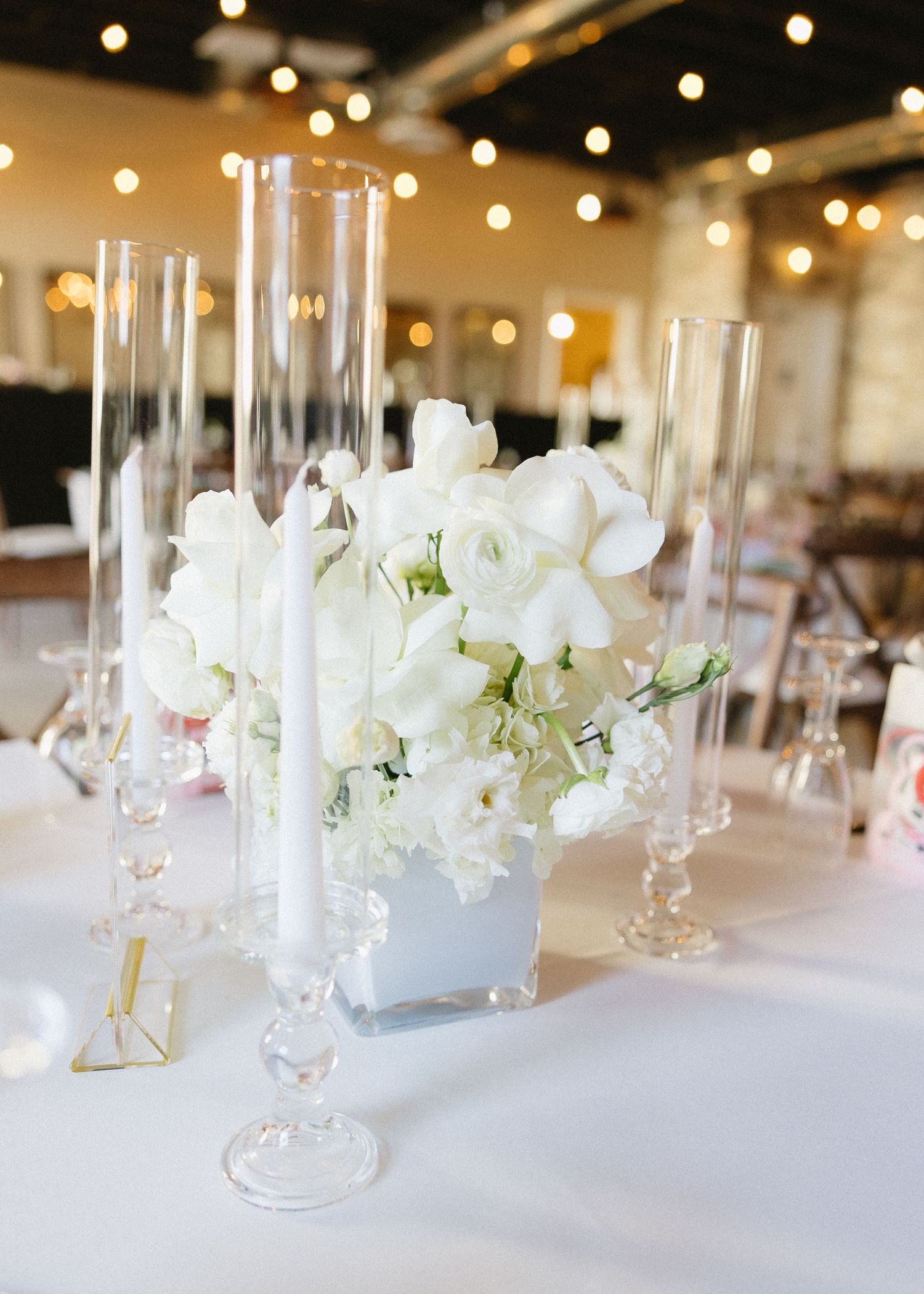 Elegant wedding table centerpiece with white flowers and tall glass cylinders under string lights.