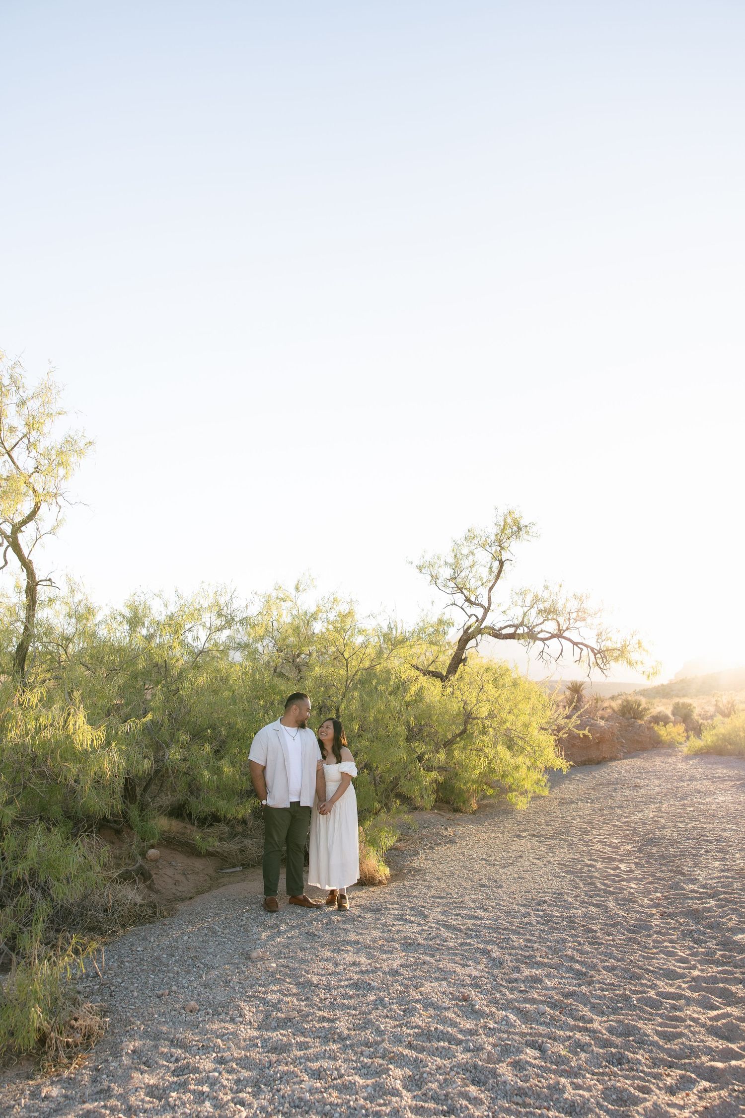 A couple stands together on a gravel path surrounded by desert vegetation at golden hour.