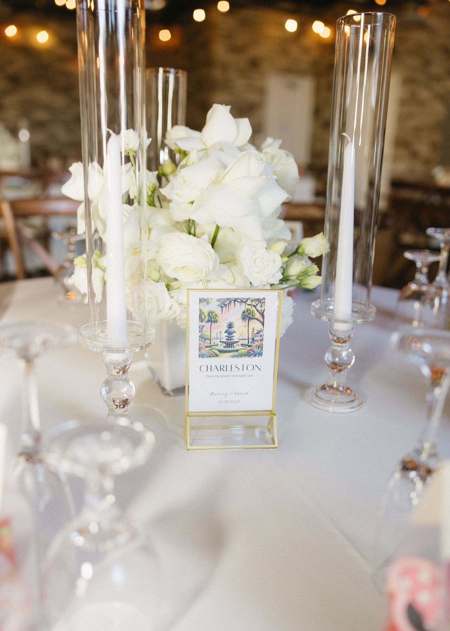 Elegant wedding table setting with white flowers, tall glass candle holders and place card on white tablecloth.