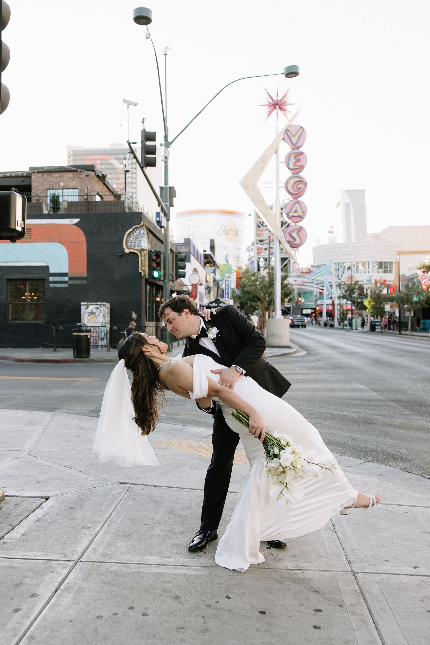 A couple in wedding attire performs a dramatic dip pose on a city street corner.