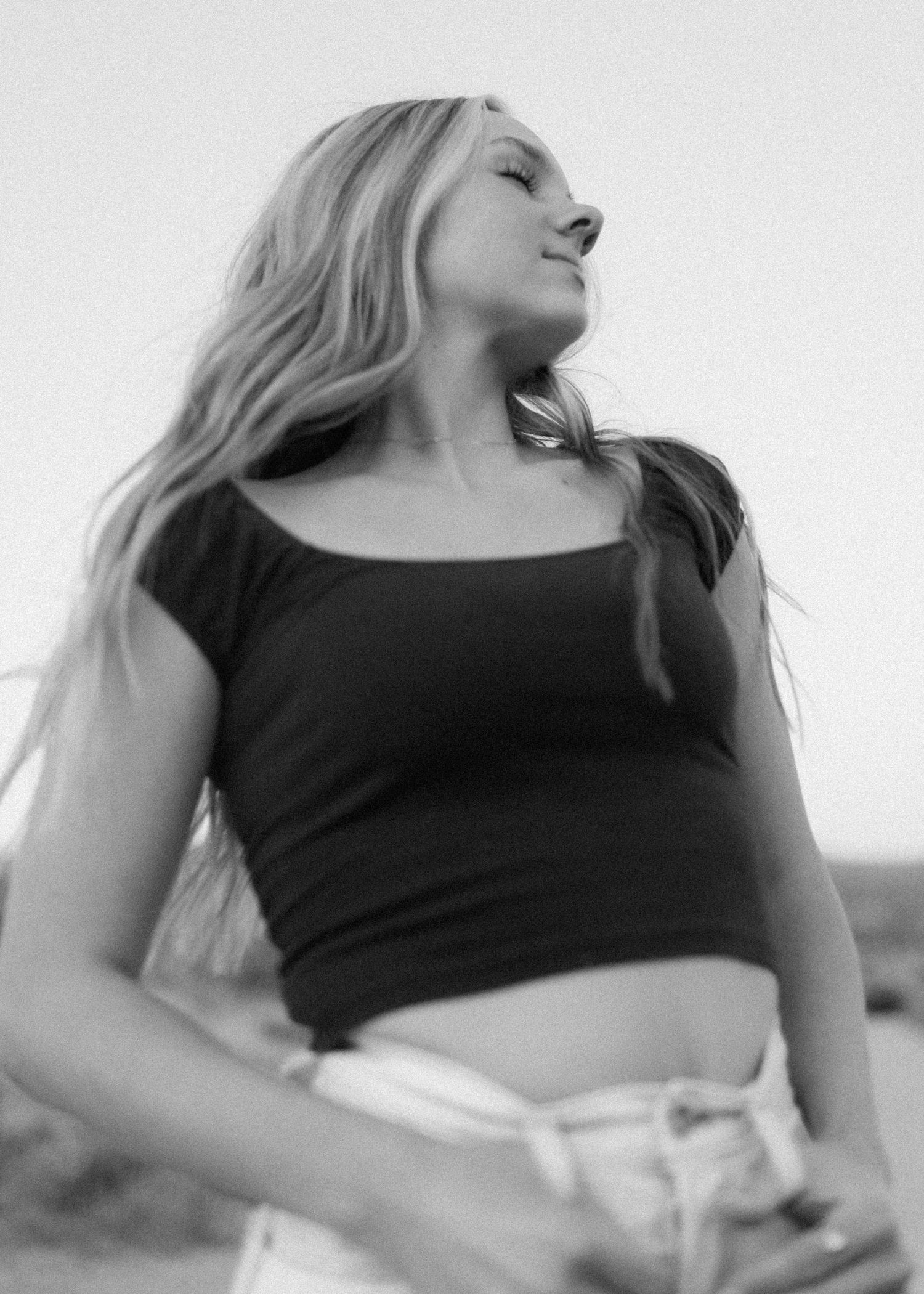 Artistic black and white portrait of a woman in casual attire against a bright sky.