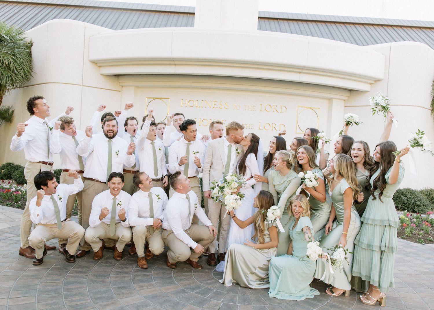 Large wedding party in light colors and pastels posed together outside venue.