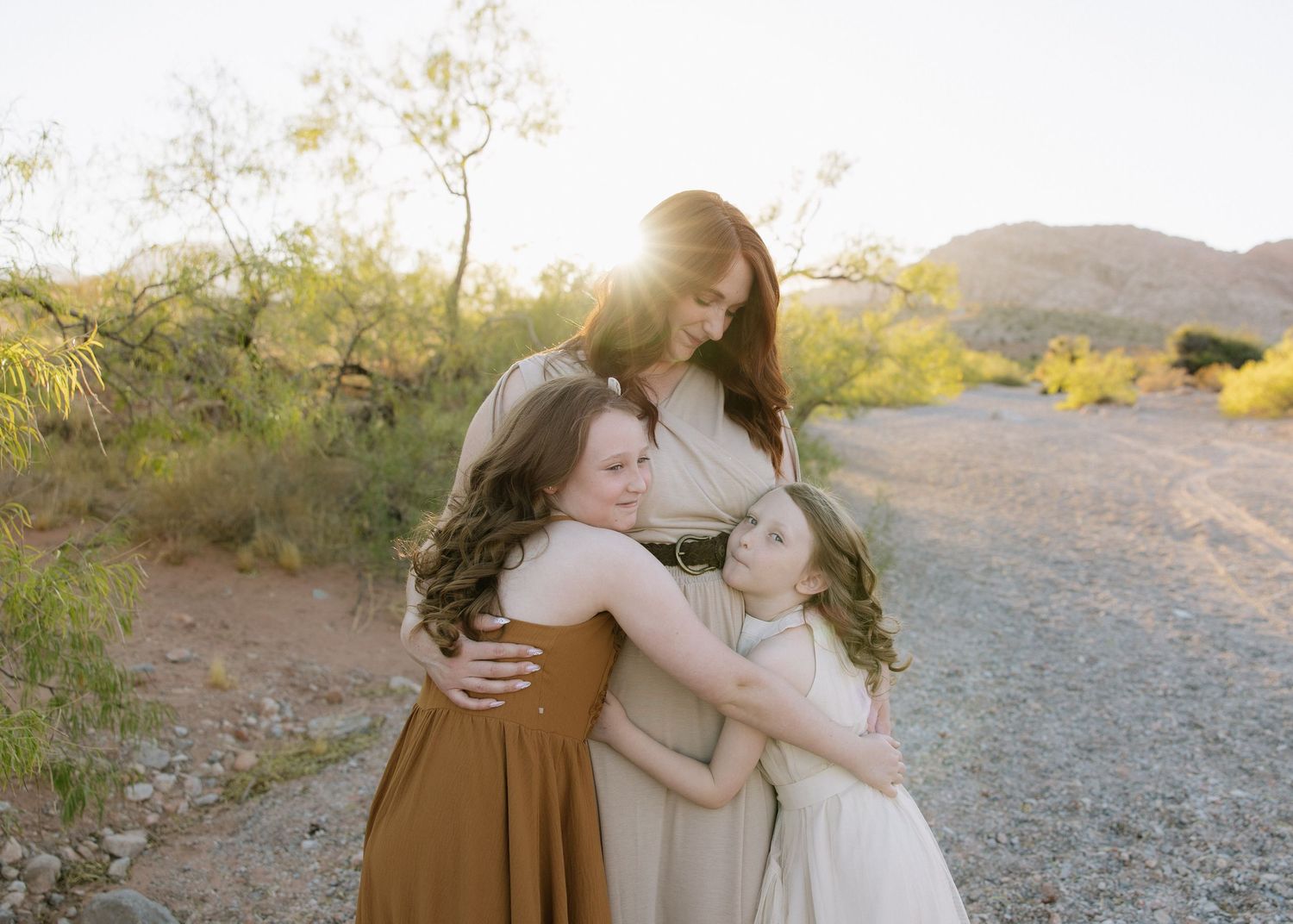 Three people embrace in a warm hug on a desert trail during golden hour.