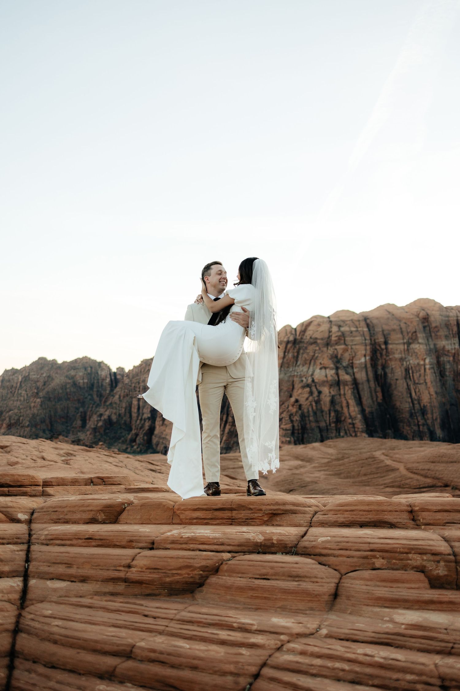 A couple in wedding attire embraces while standing on red rock formations in a desert setting.
