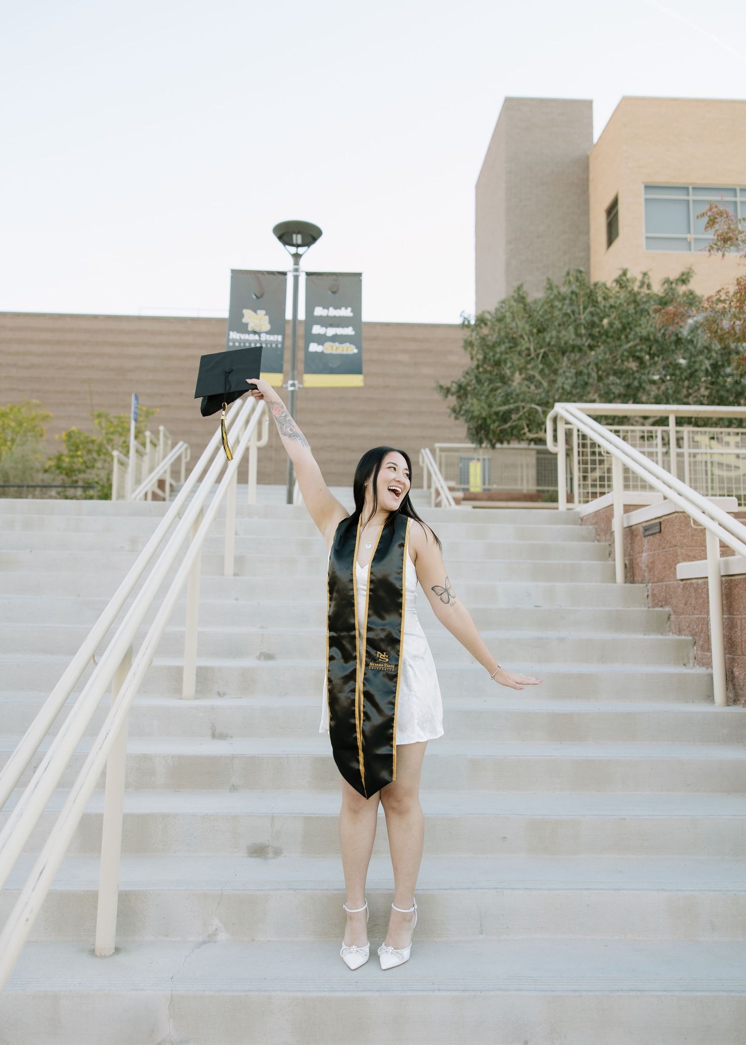 Graduate in white dress and black sash poses on campus steps.