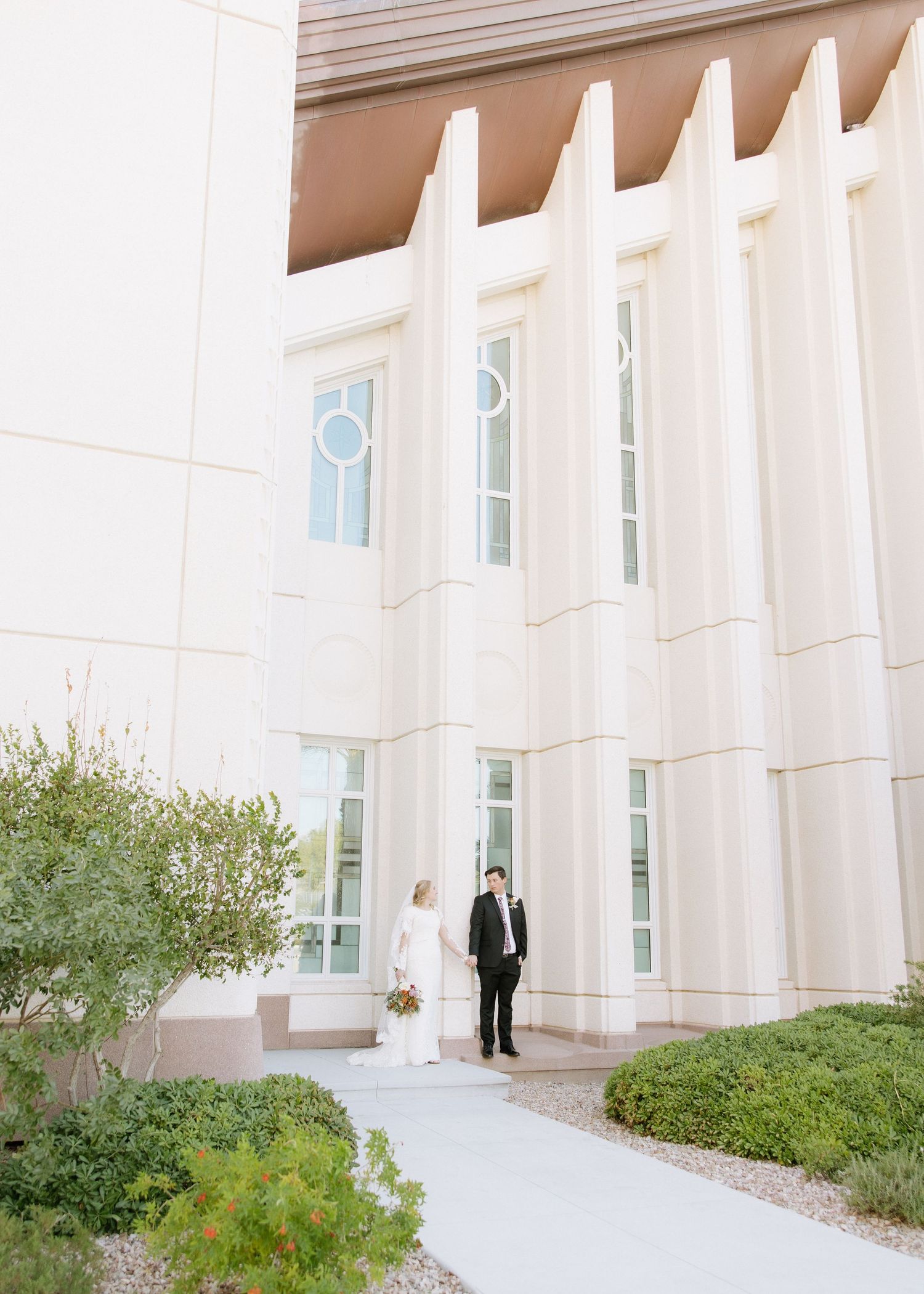 A couple stand on a white pathway in front of a modern church building with distinctive architectural columns.