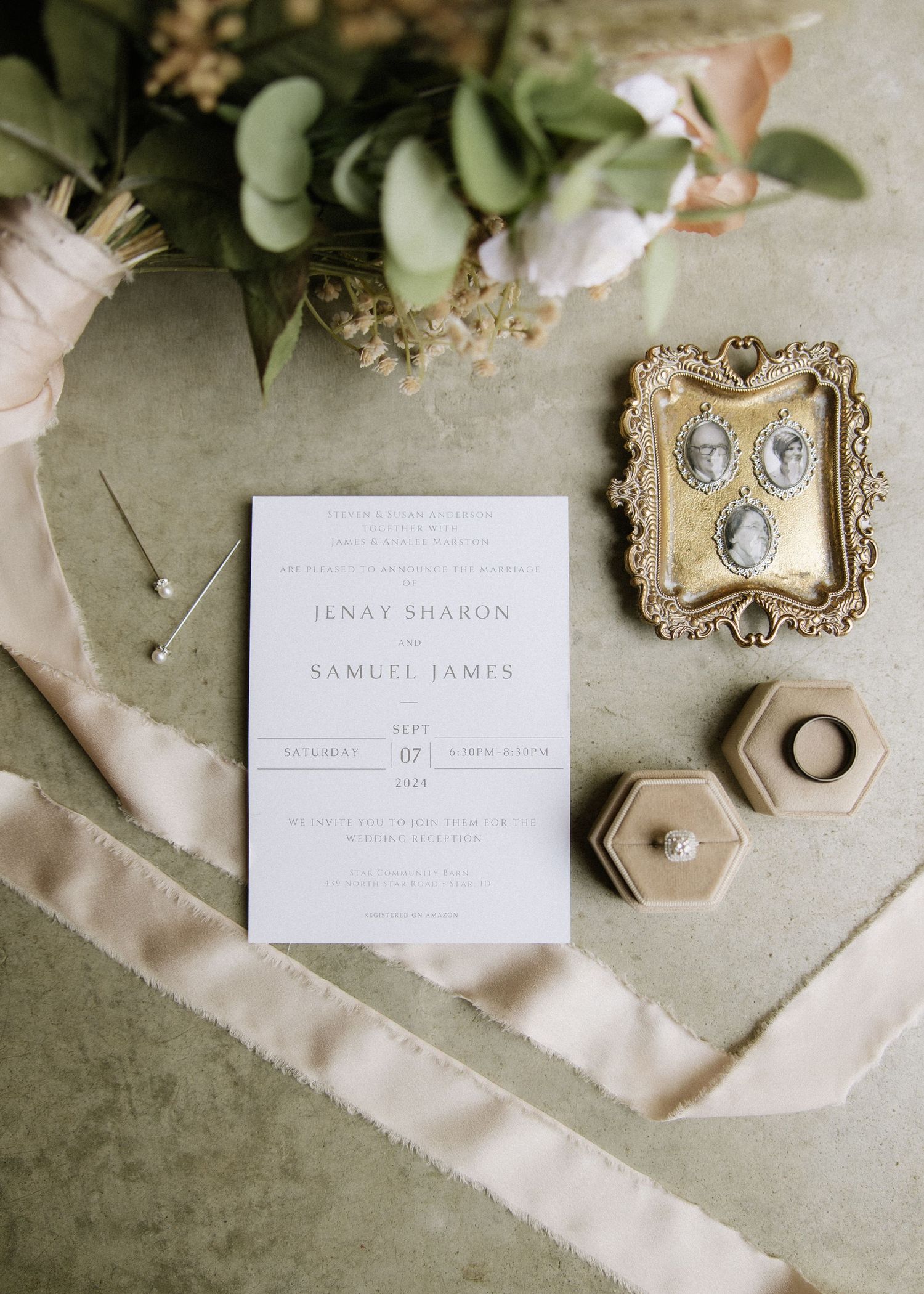 Wedding invitation and vintage gold frame displayed with greenery and ring boxes on beige fabric.