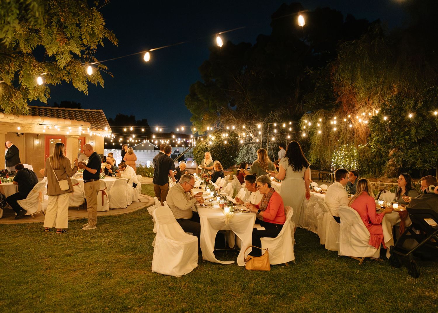Outdoor evening wedding reception with string lights illuminating tables and guests on a lawn.