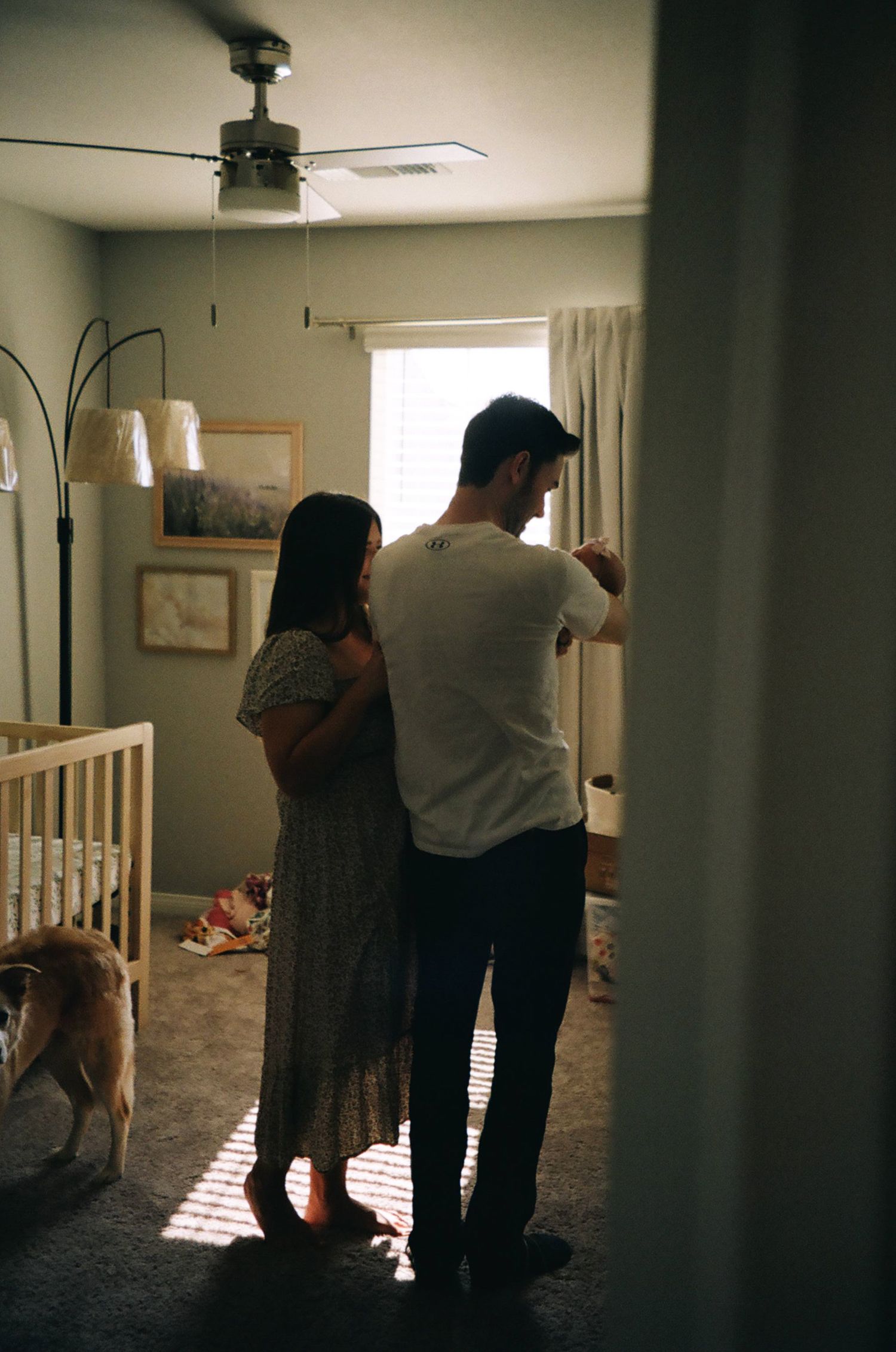 Silhouetted couple stands by window in nursery room with dog nearby.