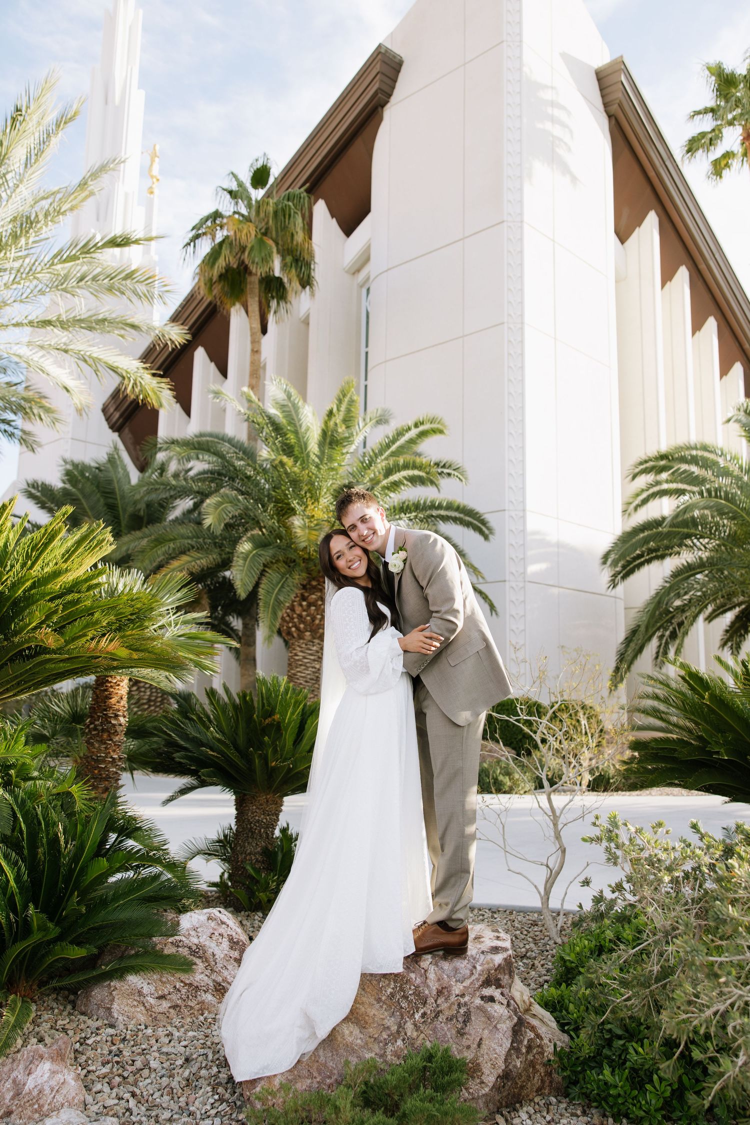 A couple shares a romantic moment among palm trees and white columns in an outdoor garden setting.