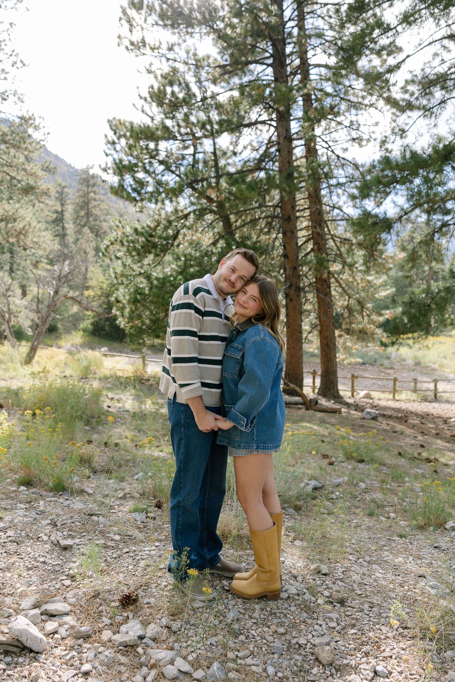 A couple embraces while standing among pine trees in a mountainous outdoor setting.
