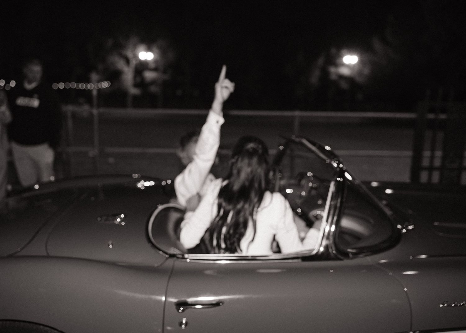Black and white photo of someone in a convertible car at night pointing upward.