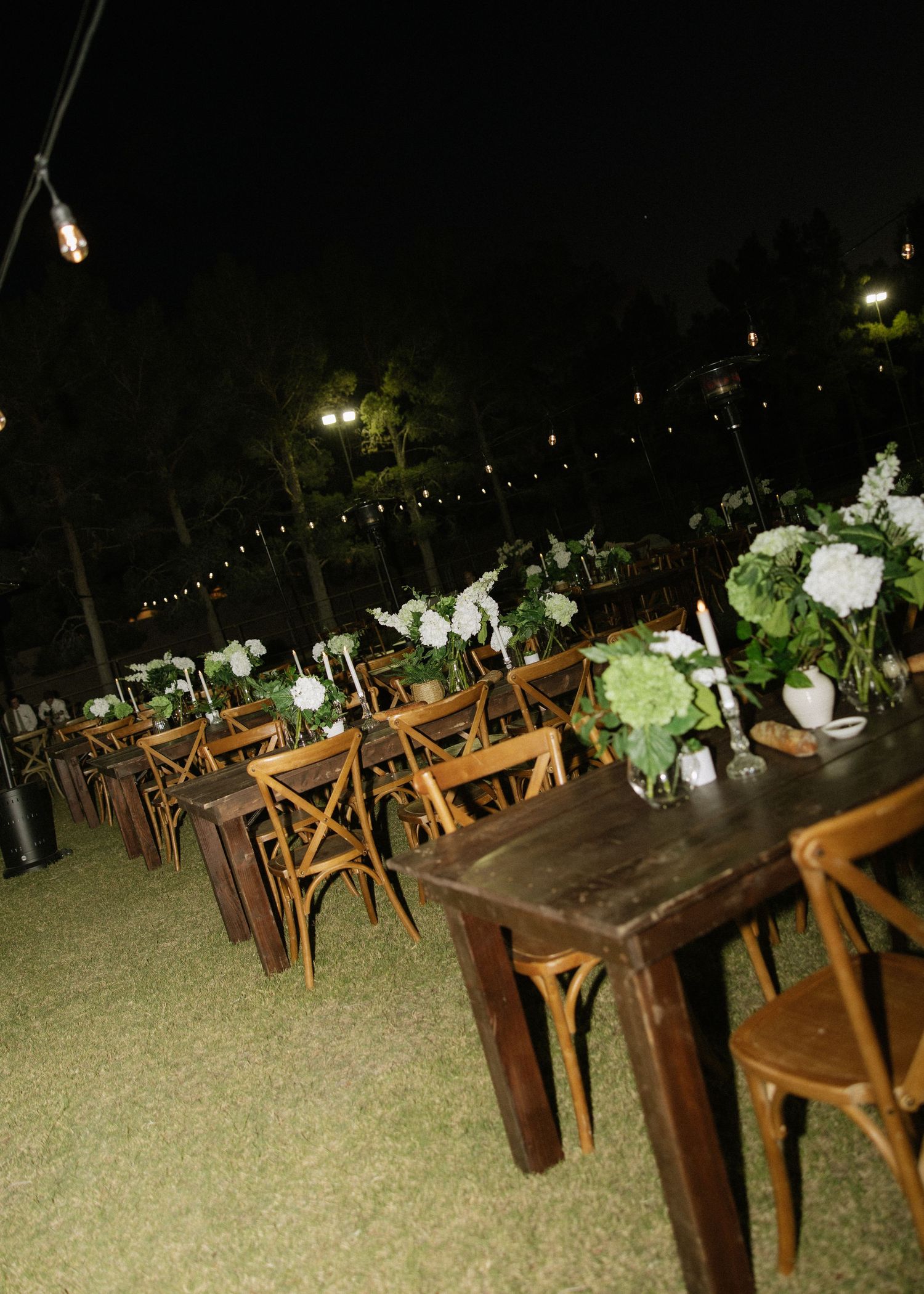 An outdoor reception area with wooden farm tables, cross-back chairs, and white floral arrangements under string lights.