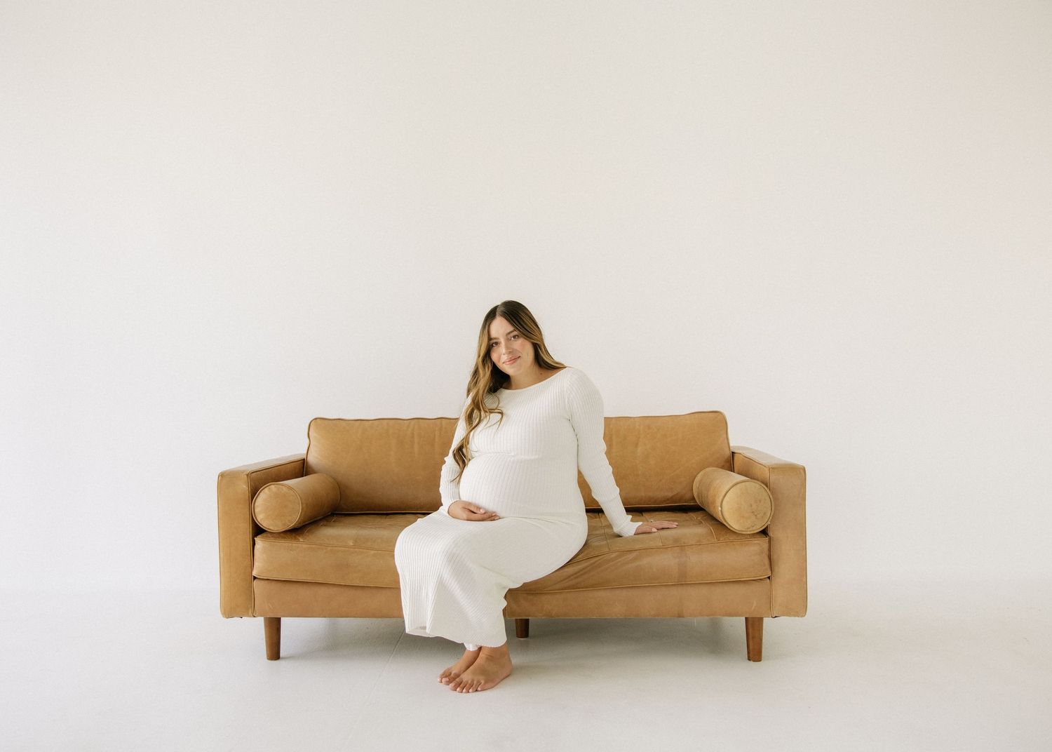 Minimalist maternity photo with woman in white dress sitting on tan couch.