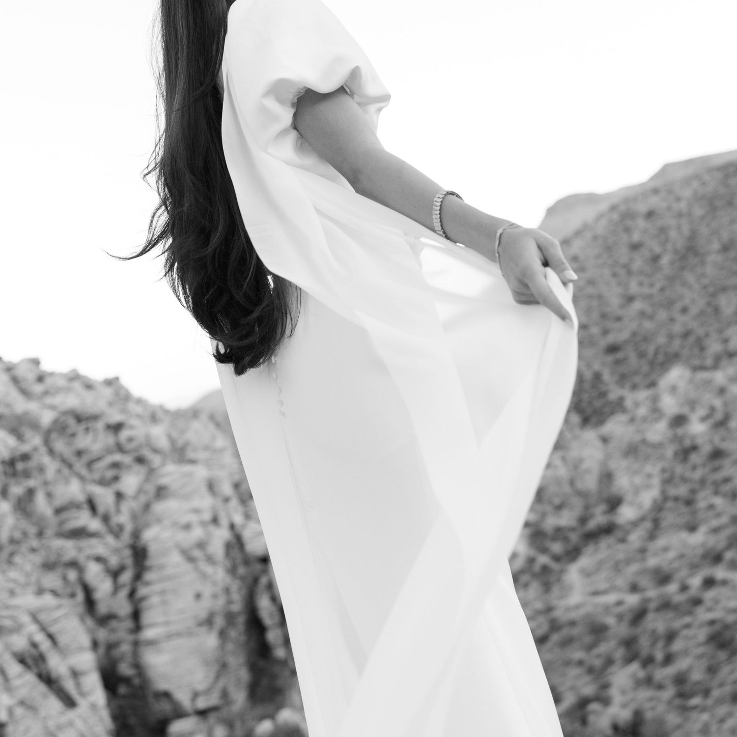A person in a flowing white dress poses gracefully against a rocky desert landscape in black and white photography.
