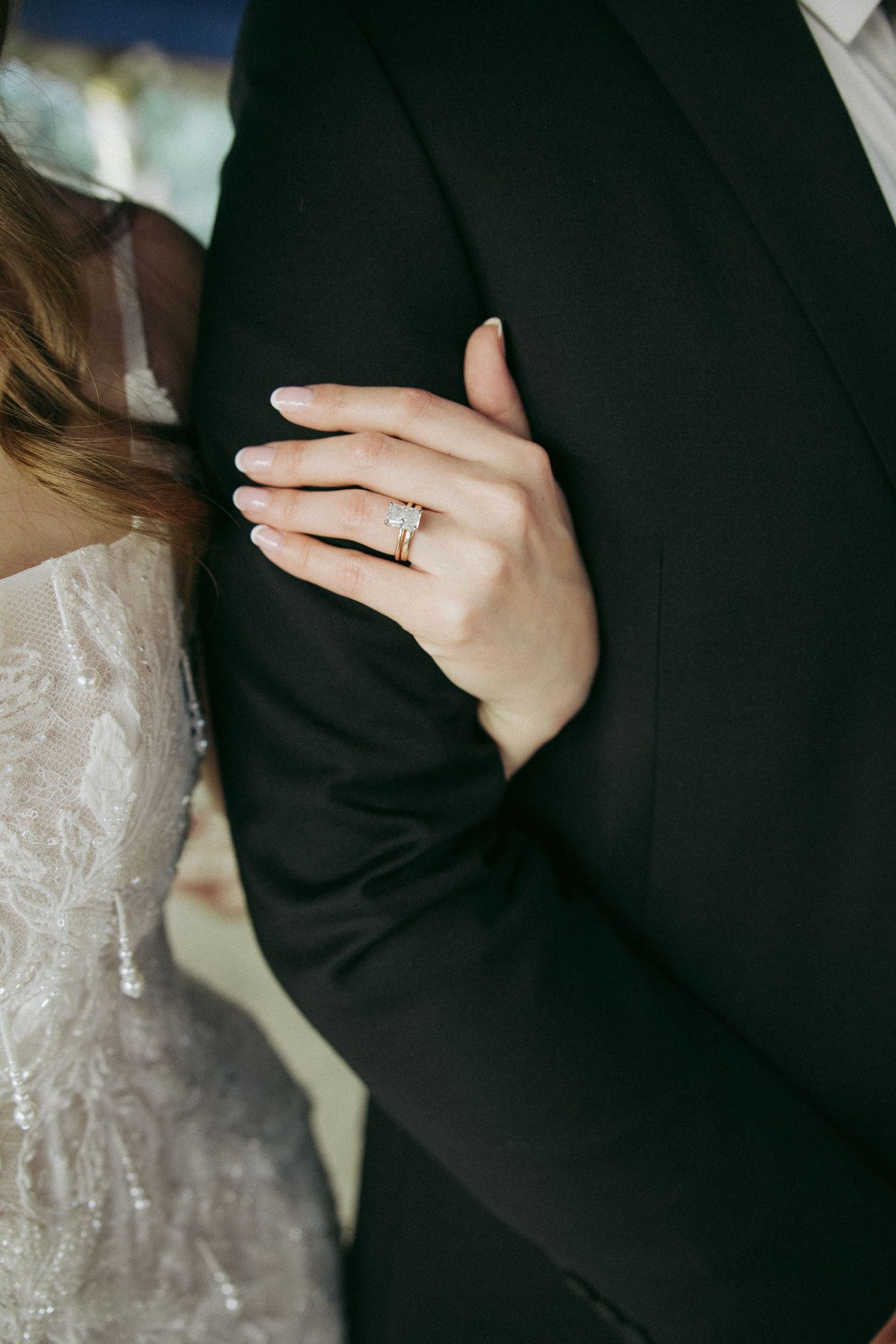 Close-up details of a couple's embrace showing white lace wedding dress against black suit with diamond ring.