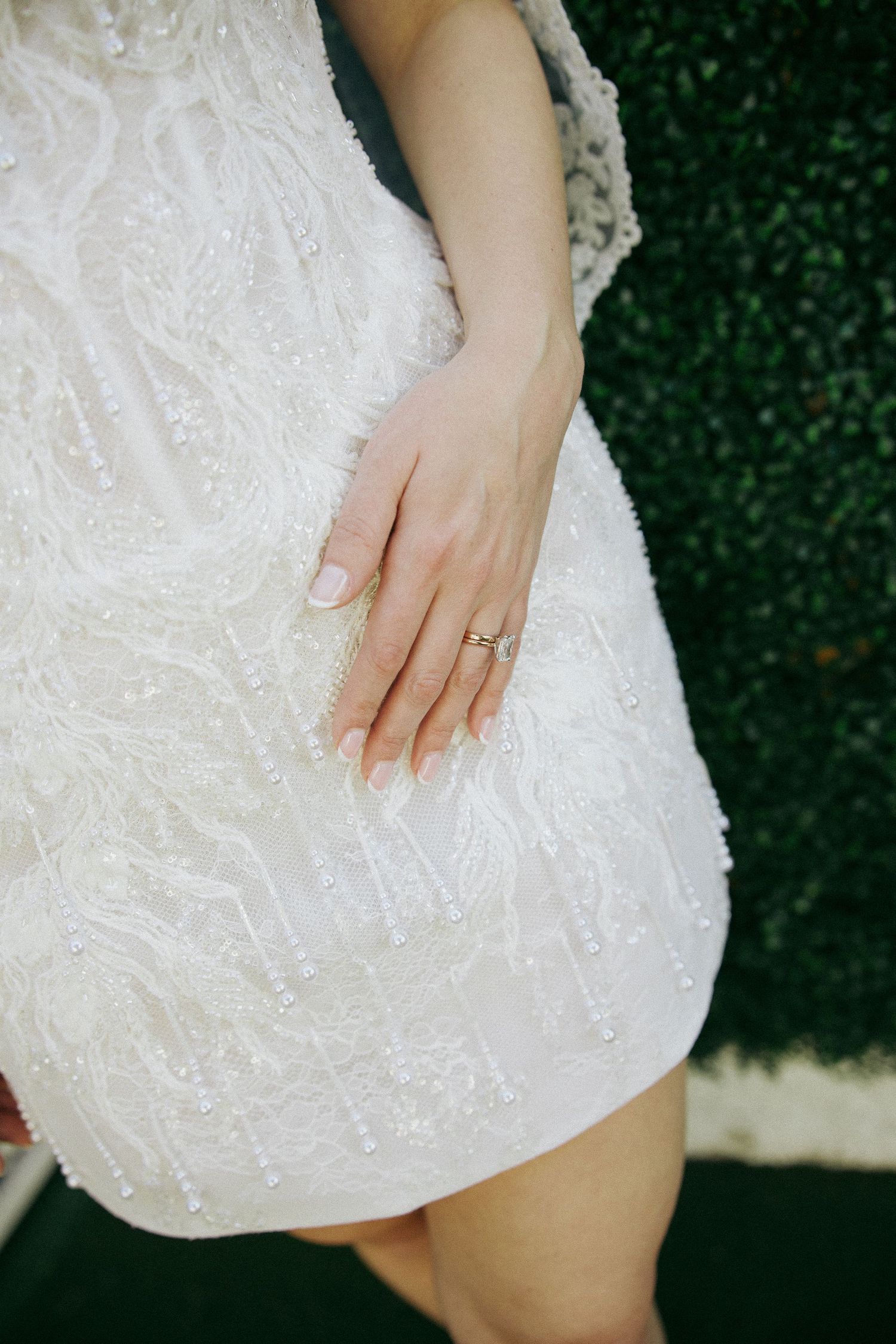 A delicate hand rests on a white beaded wedding dress against a dark green background, showcasing an engagement ring.