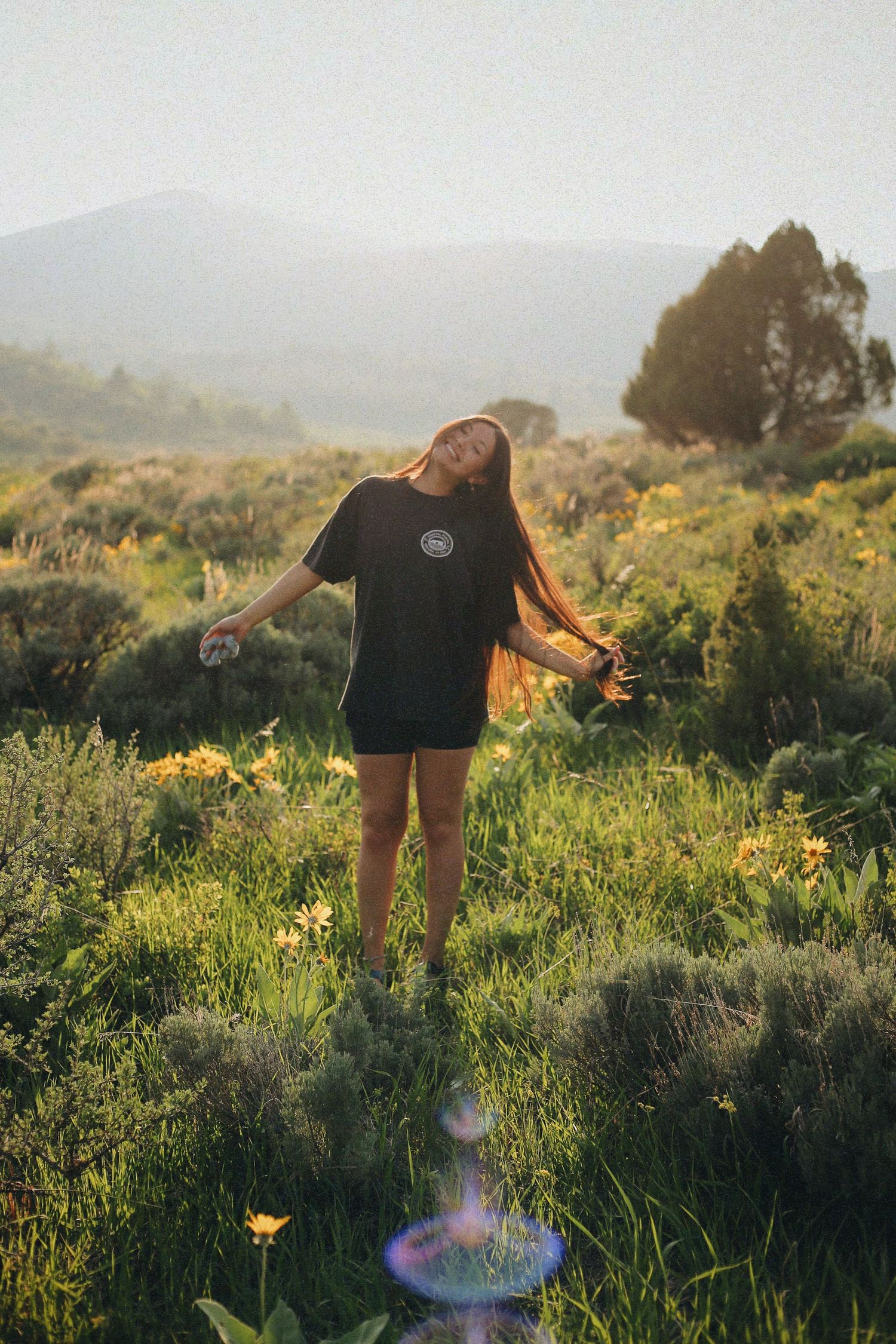 A person in a black t-shirt stands joyfully among wildflowers in a mountain meadow at sunset.