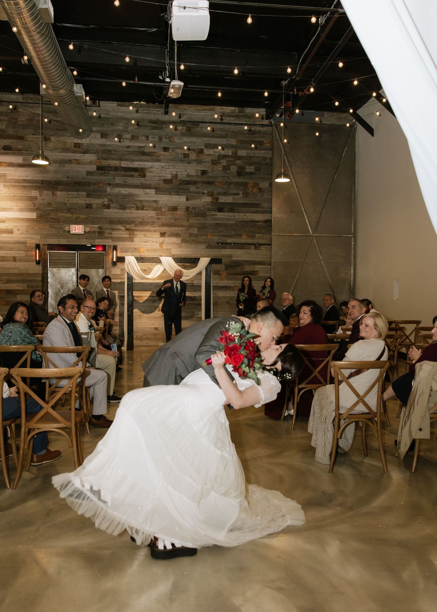 Newlyweds share a romantic dip during their first dance at a rustic venue with string lights.