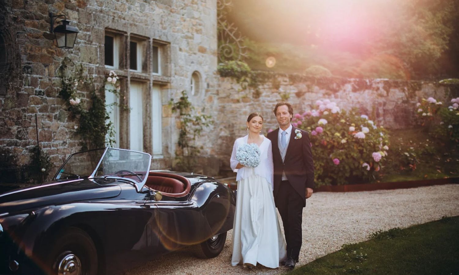 A couple poses for romantic sunset wedding photos against a rustic stone wall at their outdoor ceremony.