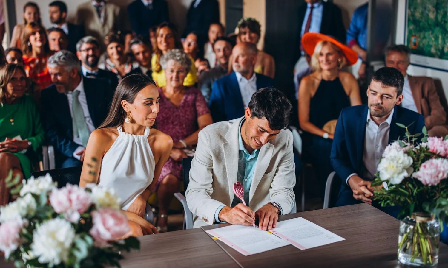A couple signs their marriage license at a wedding ceremony surrounded by family and friends.