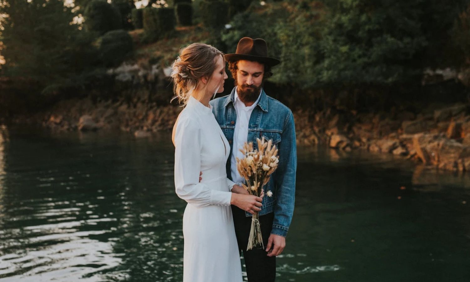 A couple shares a romantic moment by a serene lake during sunset in a scenic outdoor setting.