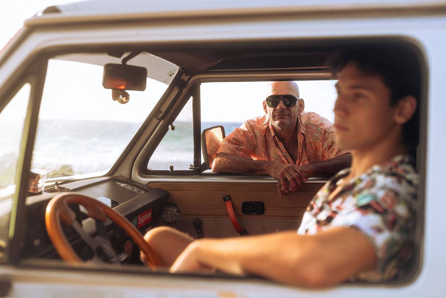 Two men in Hawaiian shirts sit in a vintage van near a sunny beach, one driving and one leaning on the door.
