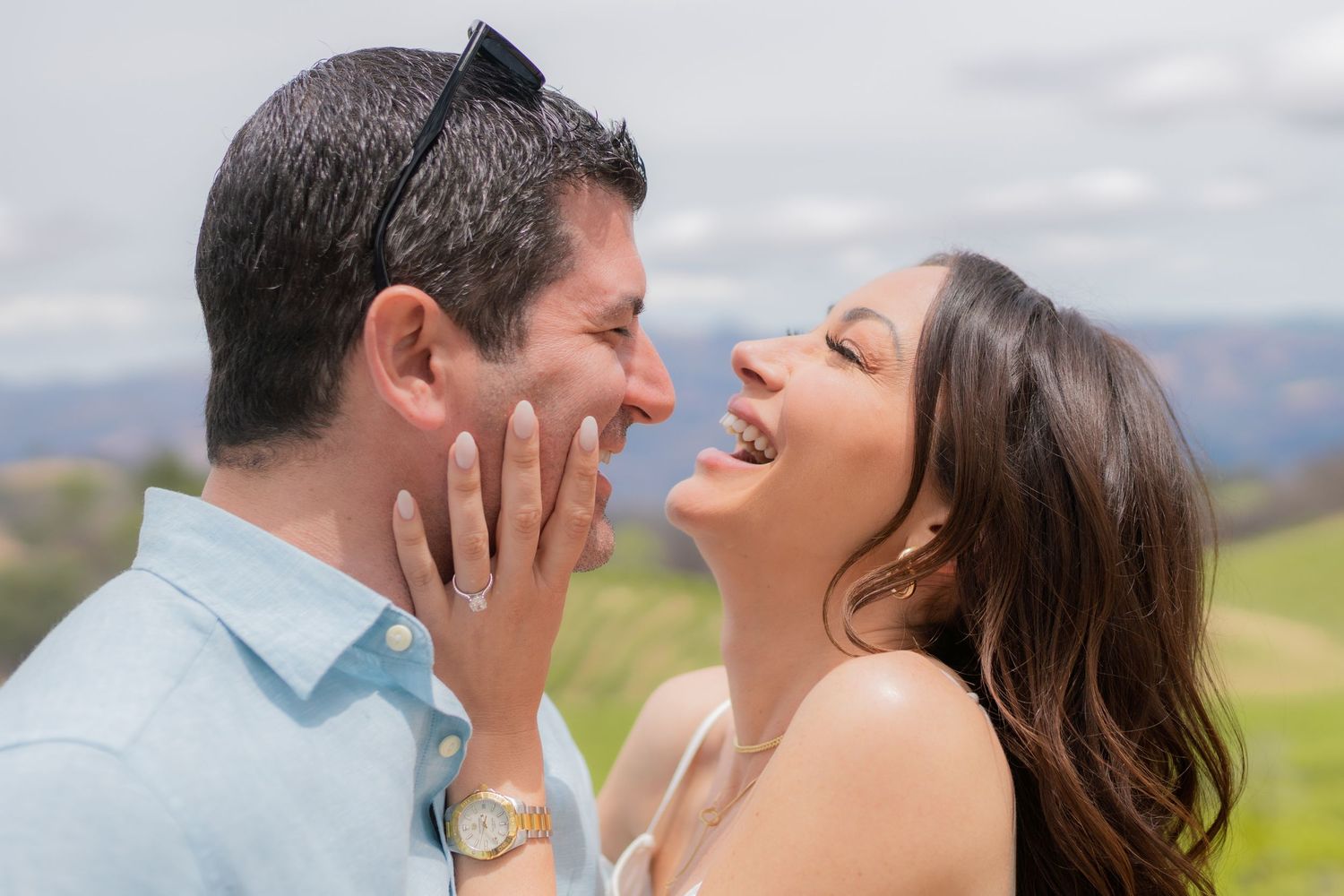 Joyful couple laughing together outdoors during engagement, woman showing diamond ring, green hills in background.