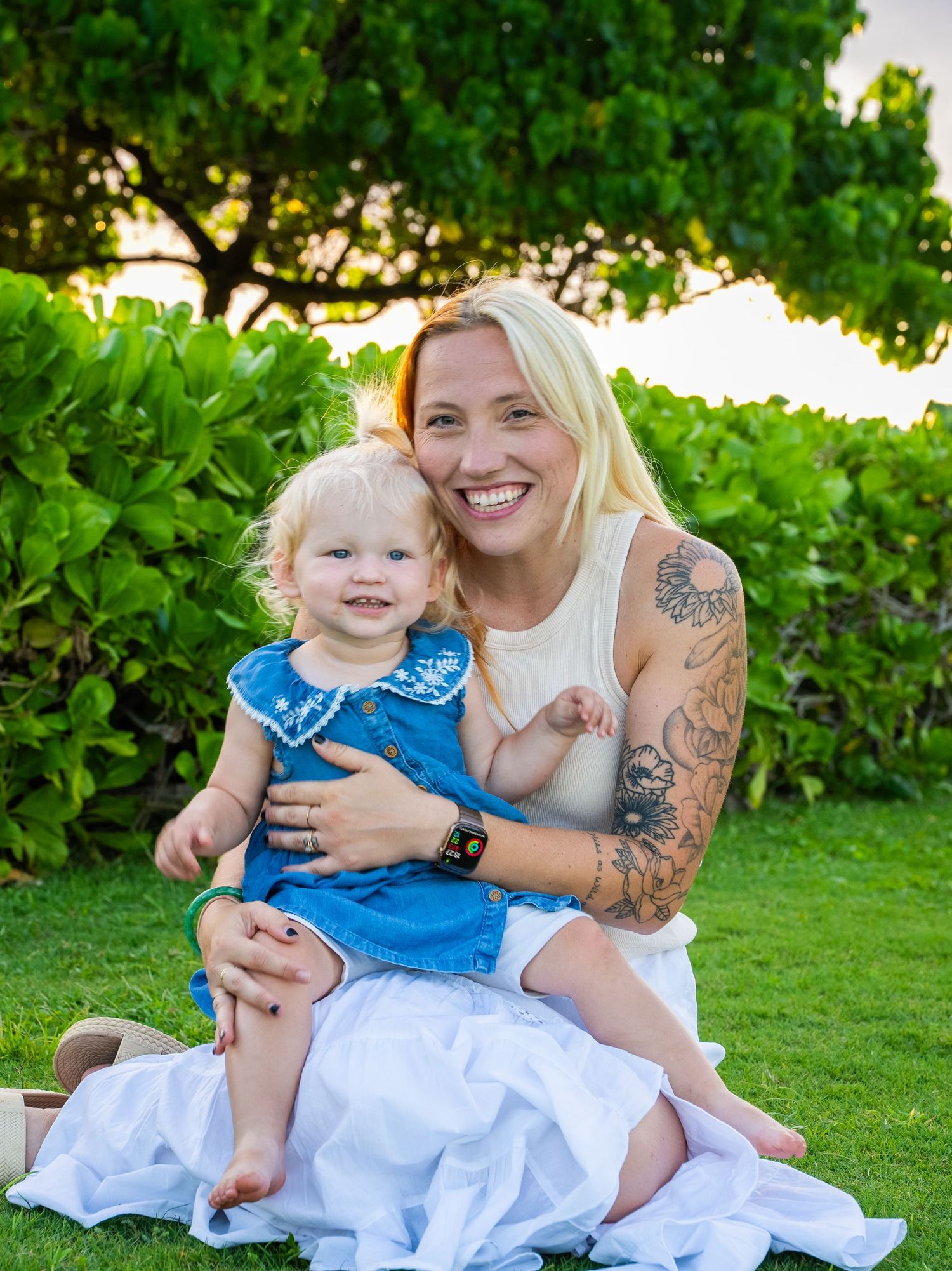 A smiling blonde woman with tattoos sits on grass holding a toddler girl in a blue dress outdoors.