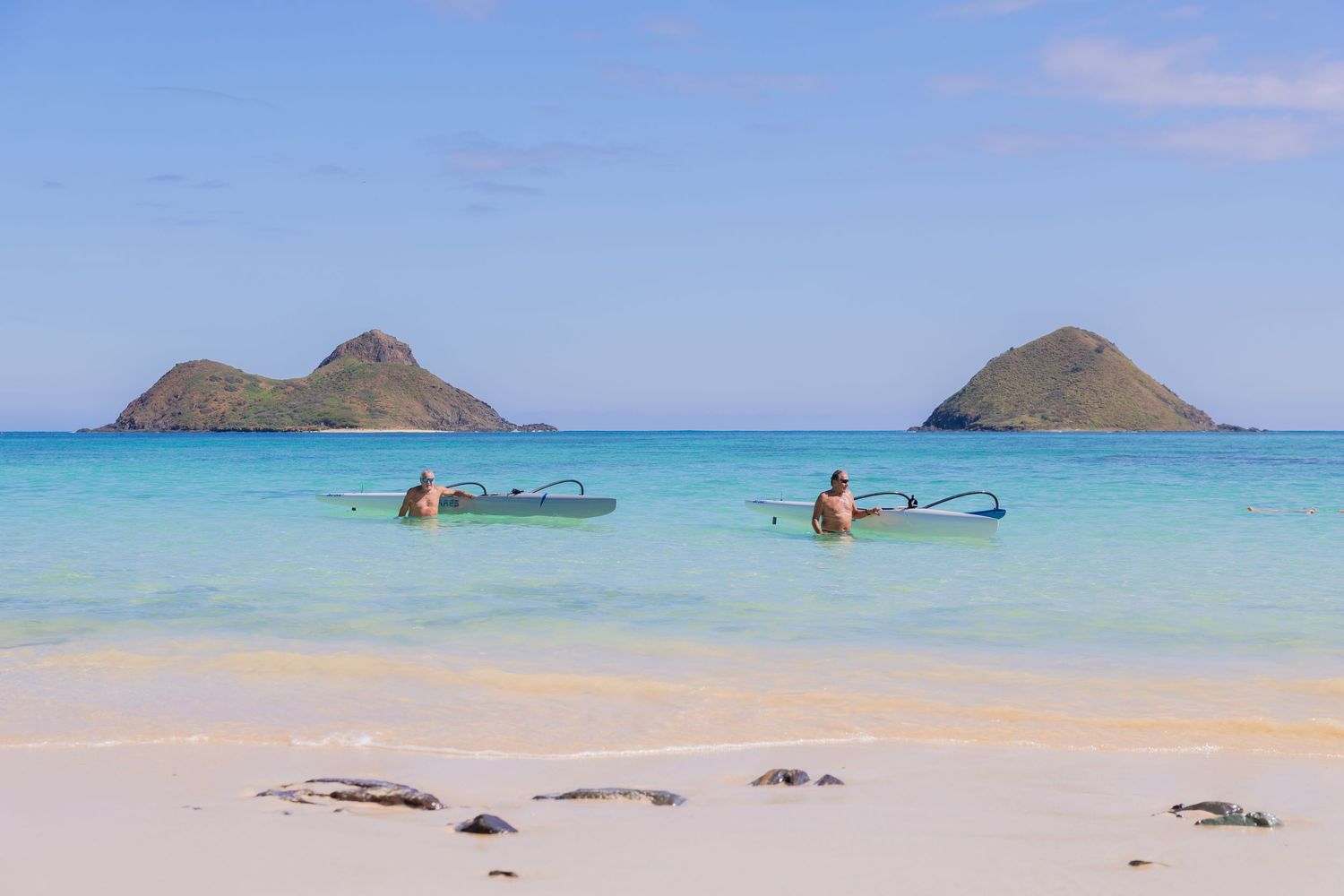Two kayakers paddle in crystal-clear turquoise waters near Lanikai Beach, Hawaii, with twin Mokulua Islands in the background.