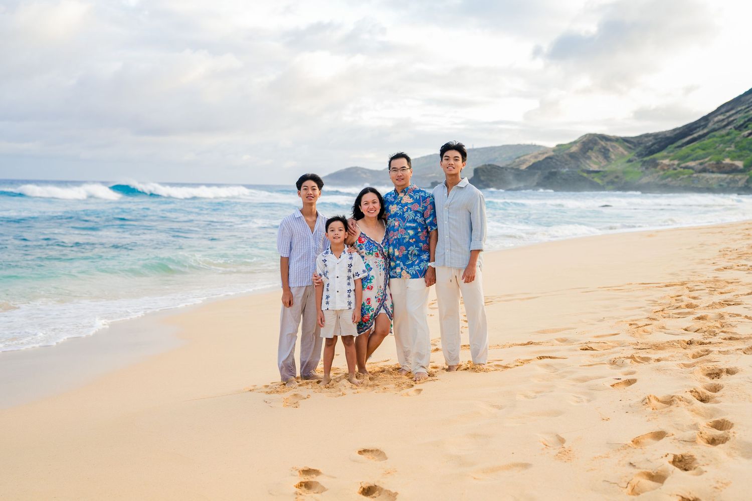 Family of five posing together on a sandy Hawaiian beach with turquoise waves and green mountains in the background.
