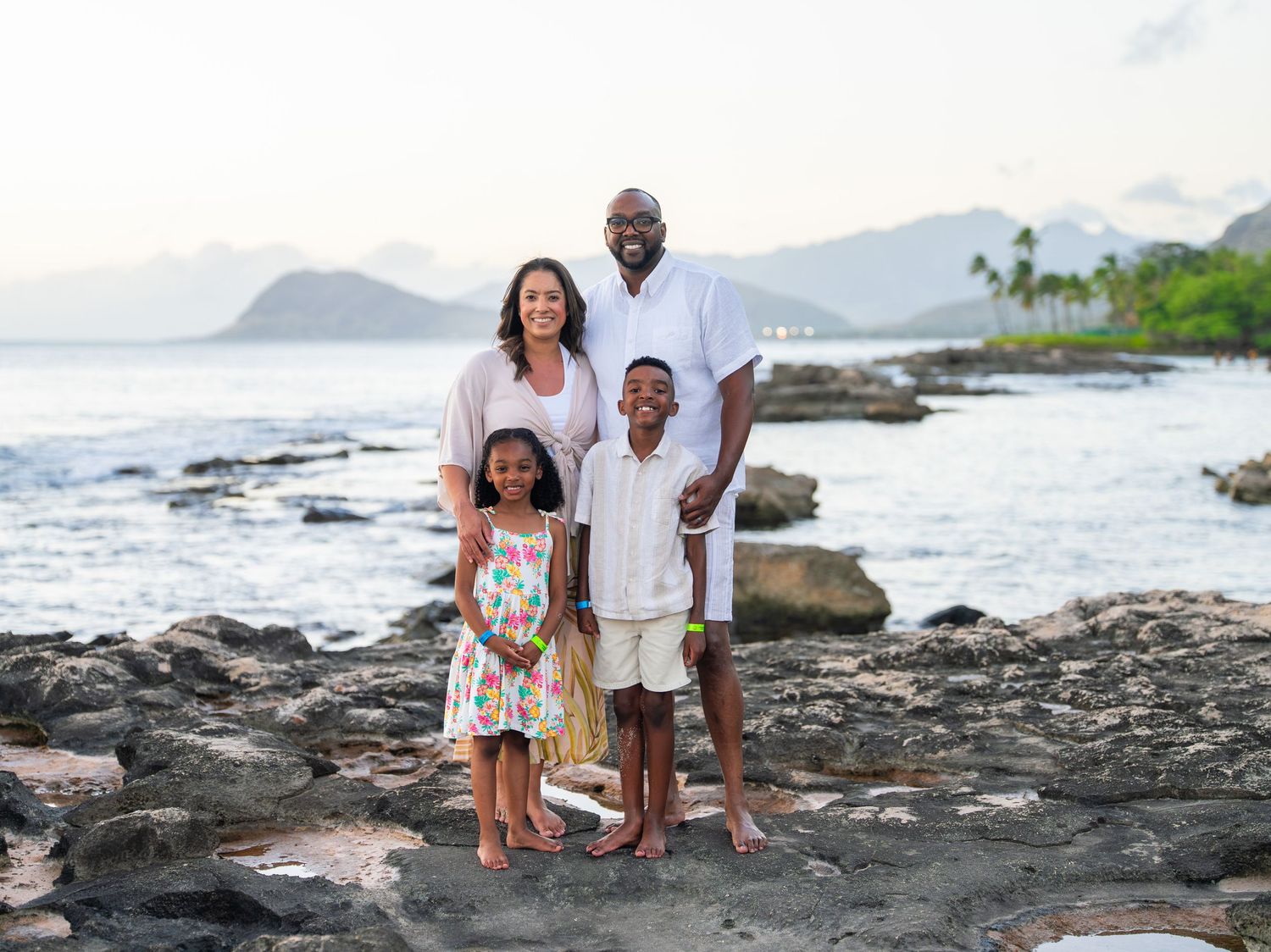 Family of four posing on rocky beach shore with ocean and mountains in background at sunset.