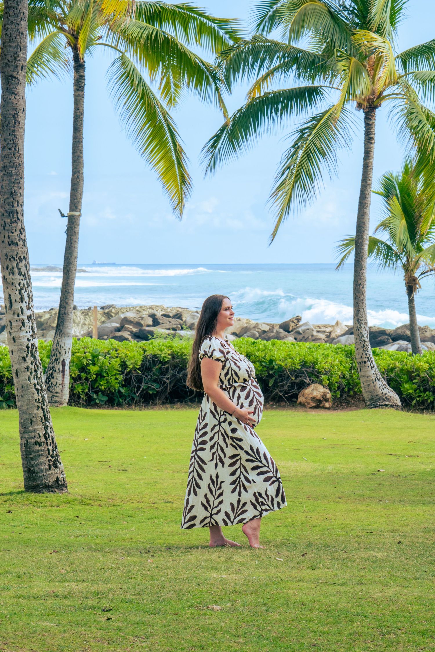 Pregnant woman in floral maxi dress standing barefoot on green lawn with tropical palm trees and ocean waves in background.