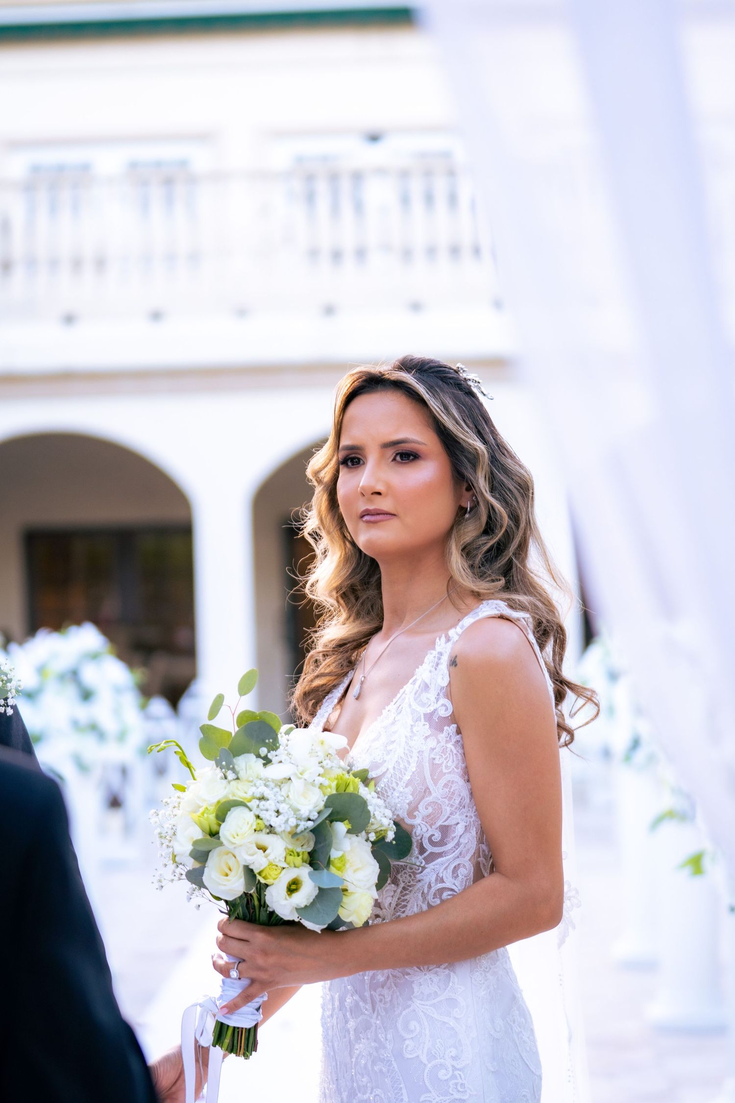 Bride in white lace gown holding white floral bouquet stands outdoors at elegant wedding venue.