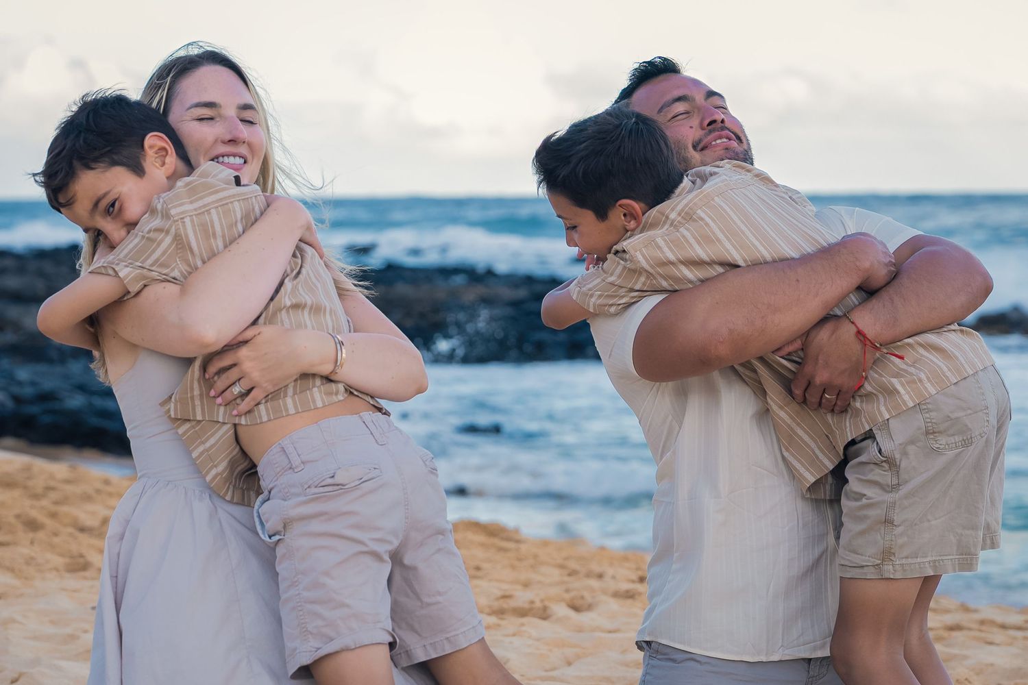 Family hugging on a sandy beach at sunset, parents embracing children in matching outfits near ocean waves.