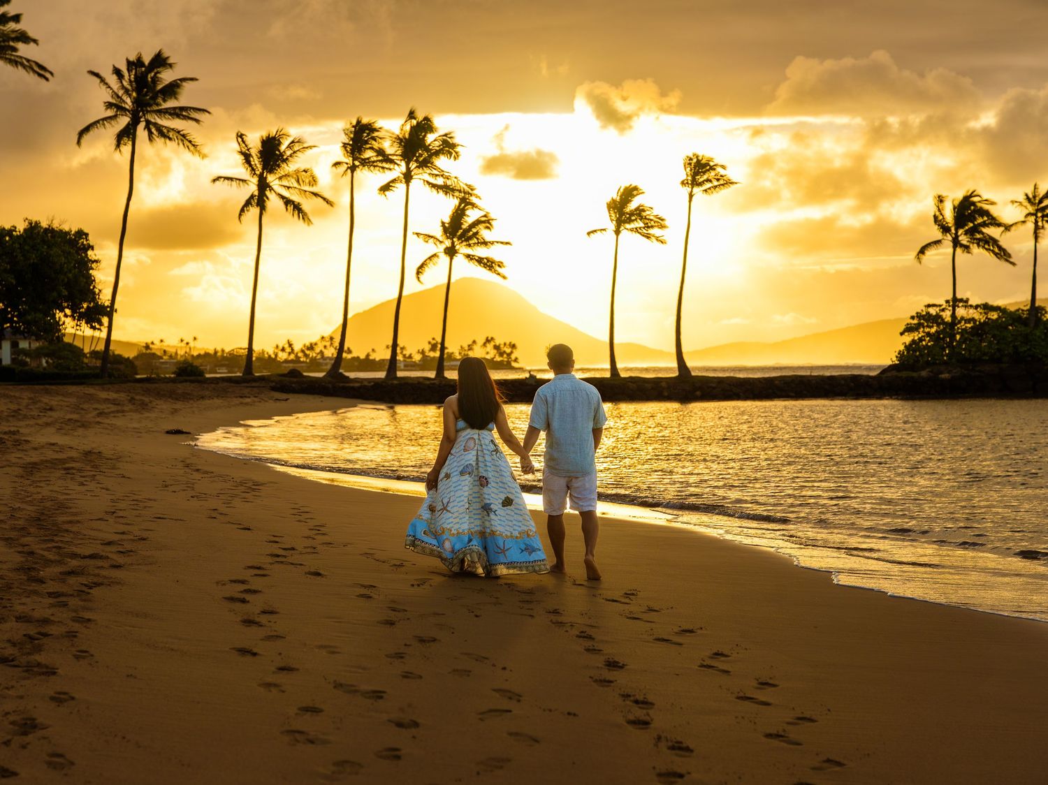 Couple holding hands walking on a tropical beach at golden sunset with palm trees and mountain views.