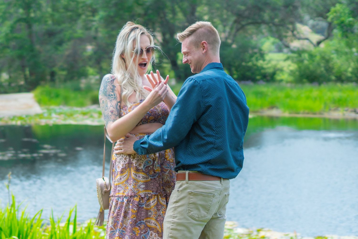 Couple sharing a romantic moment by a serene lake, woman showing engagement ring outdoors.