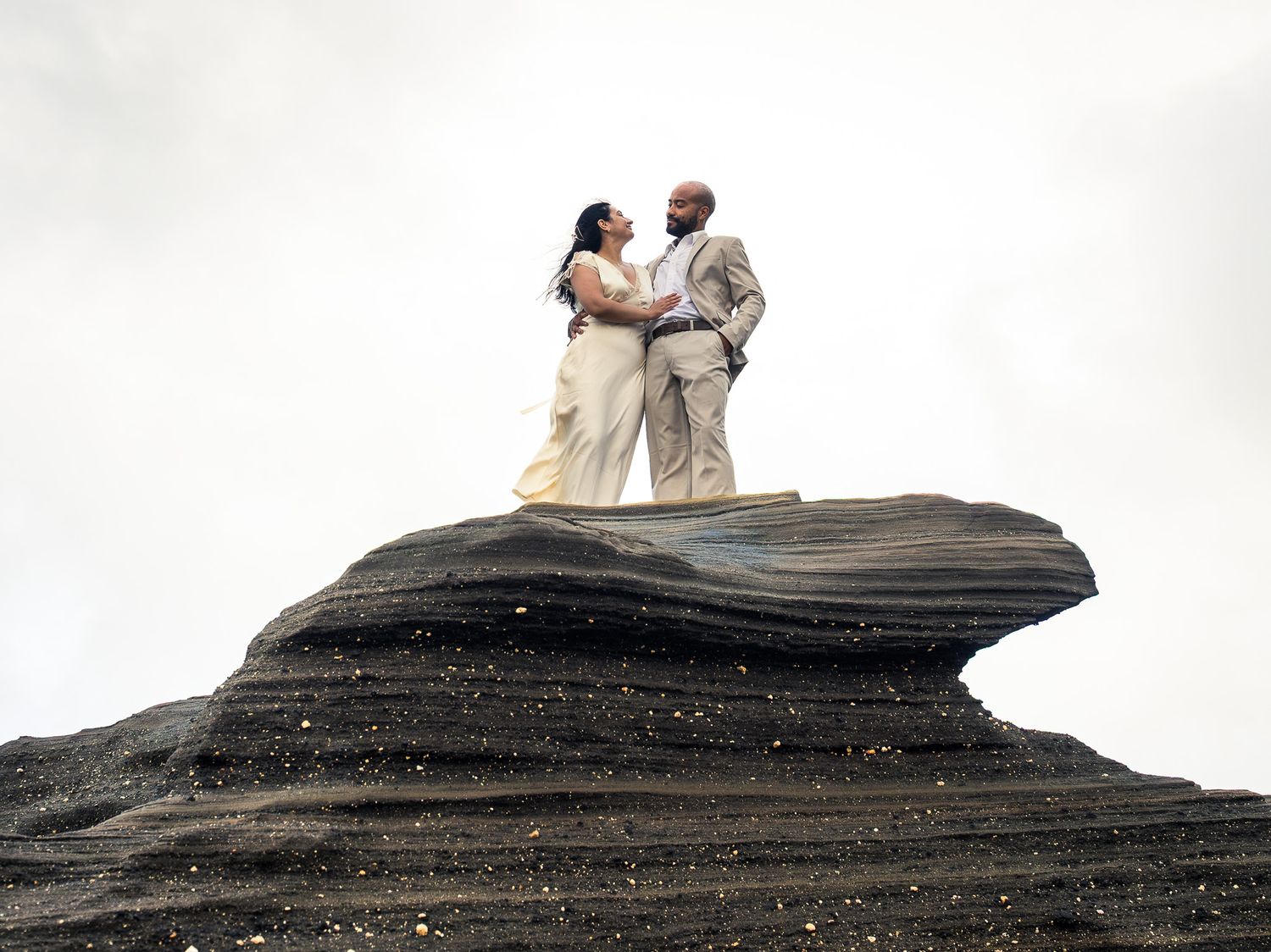 Couple in formal attire standing atop a dramatic rock formation, sharing a romantic embrace against a cloudy sky.