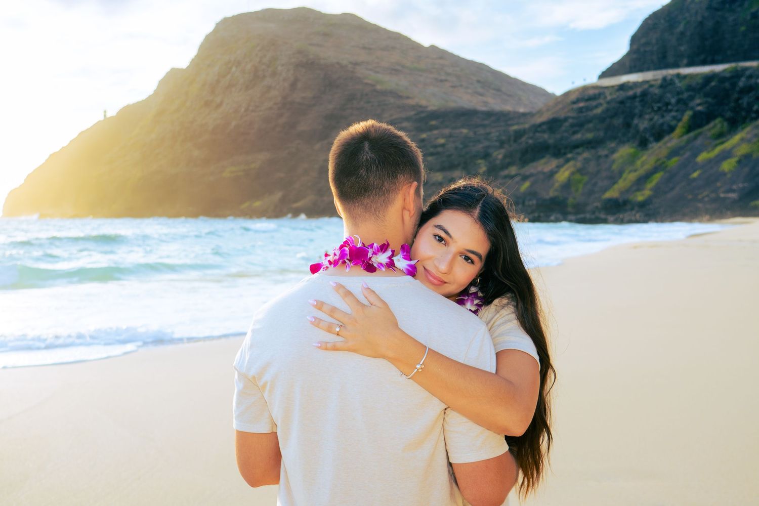 Couple embracing on a Hawaiian beach with purple lei, ocean waves and mountain cliffs in the background.