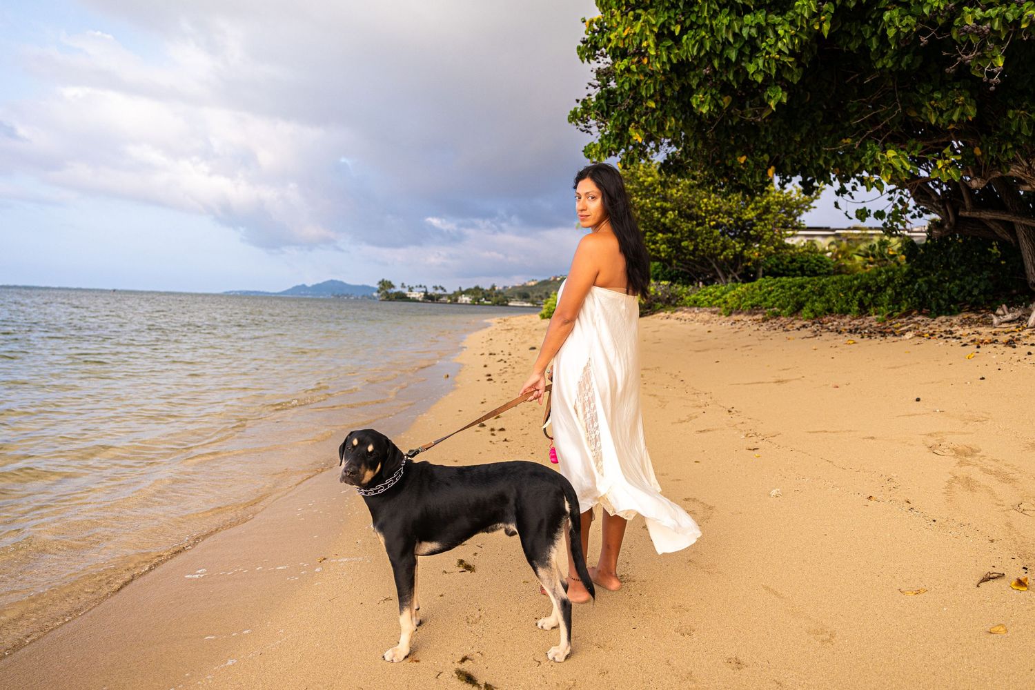 Woman in white dress walking black dog on tropical beach at sunset with calm ocean waters.