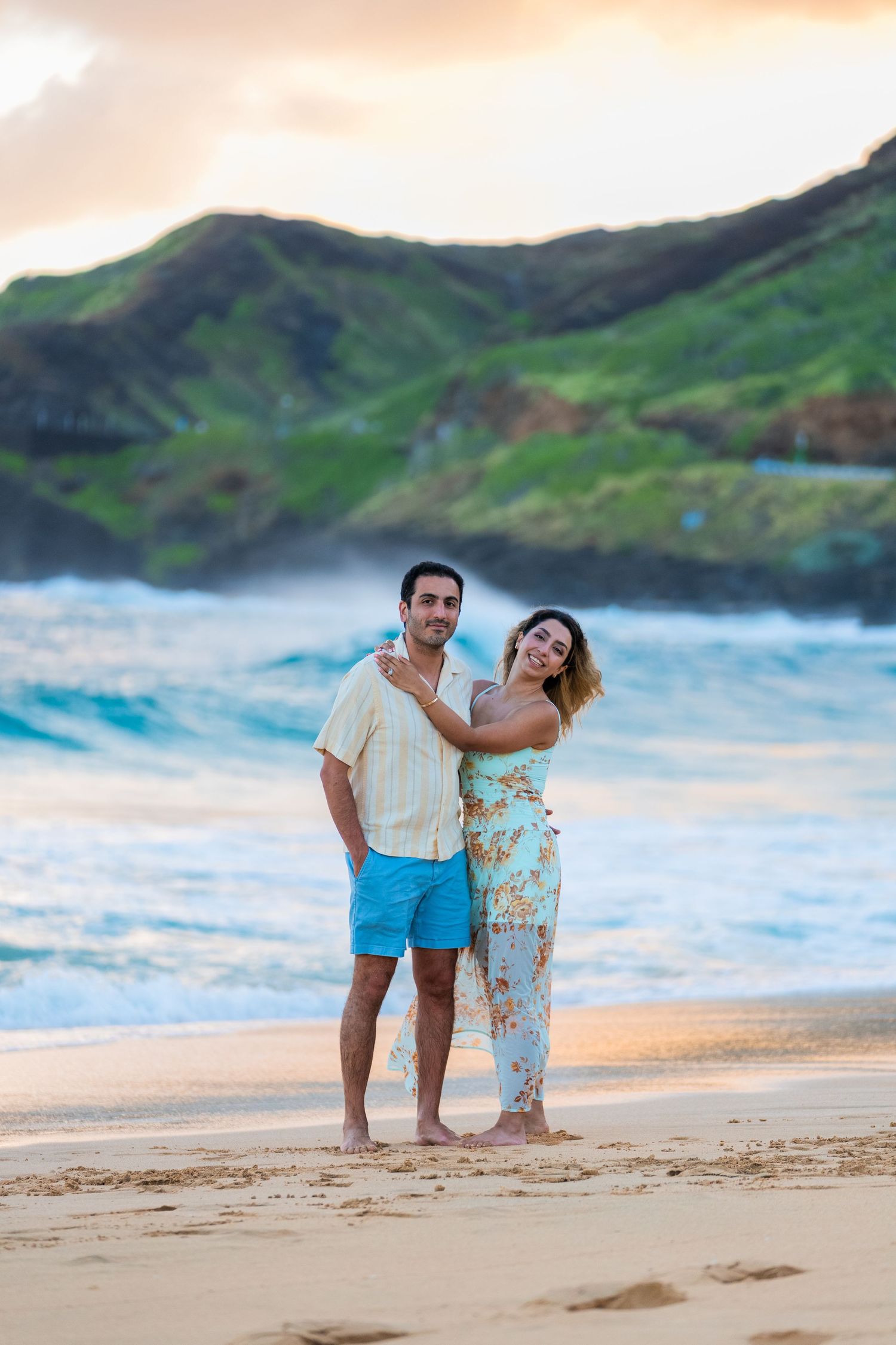 Couple embracing on a sandy beach at sunset with green mountains and ocean waves in Hawaii.