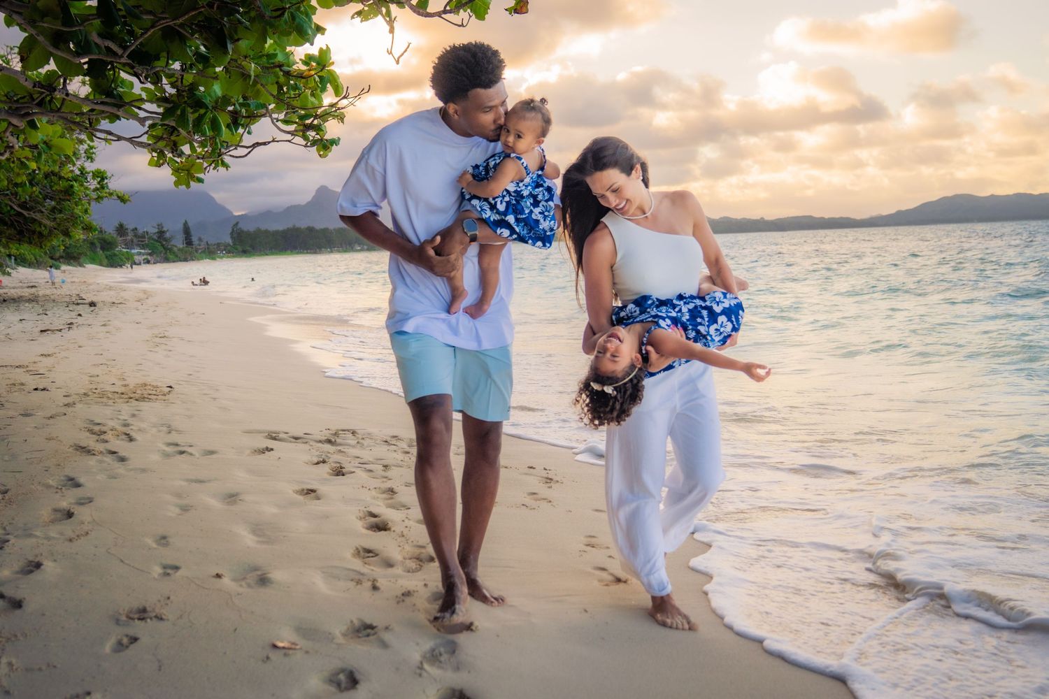 Family of four enjoying a sunset beach walk, parents playfully holding children on a tropical sandy shore.