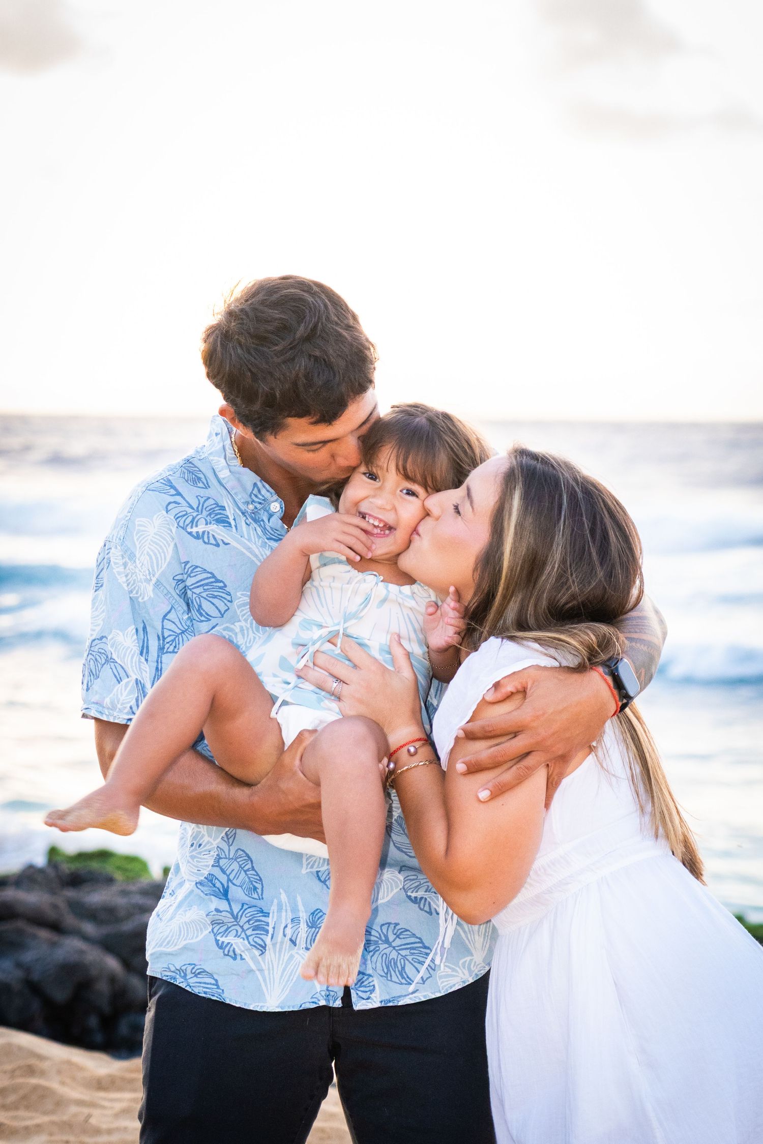 Happy family of three sharing kisses on a sandy beach at sunset, parents kissing their smiling toddler.