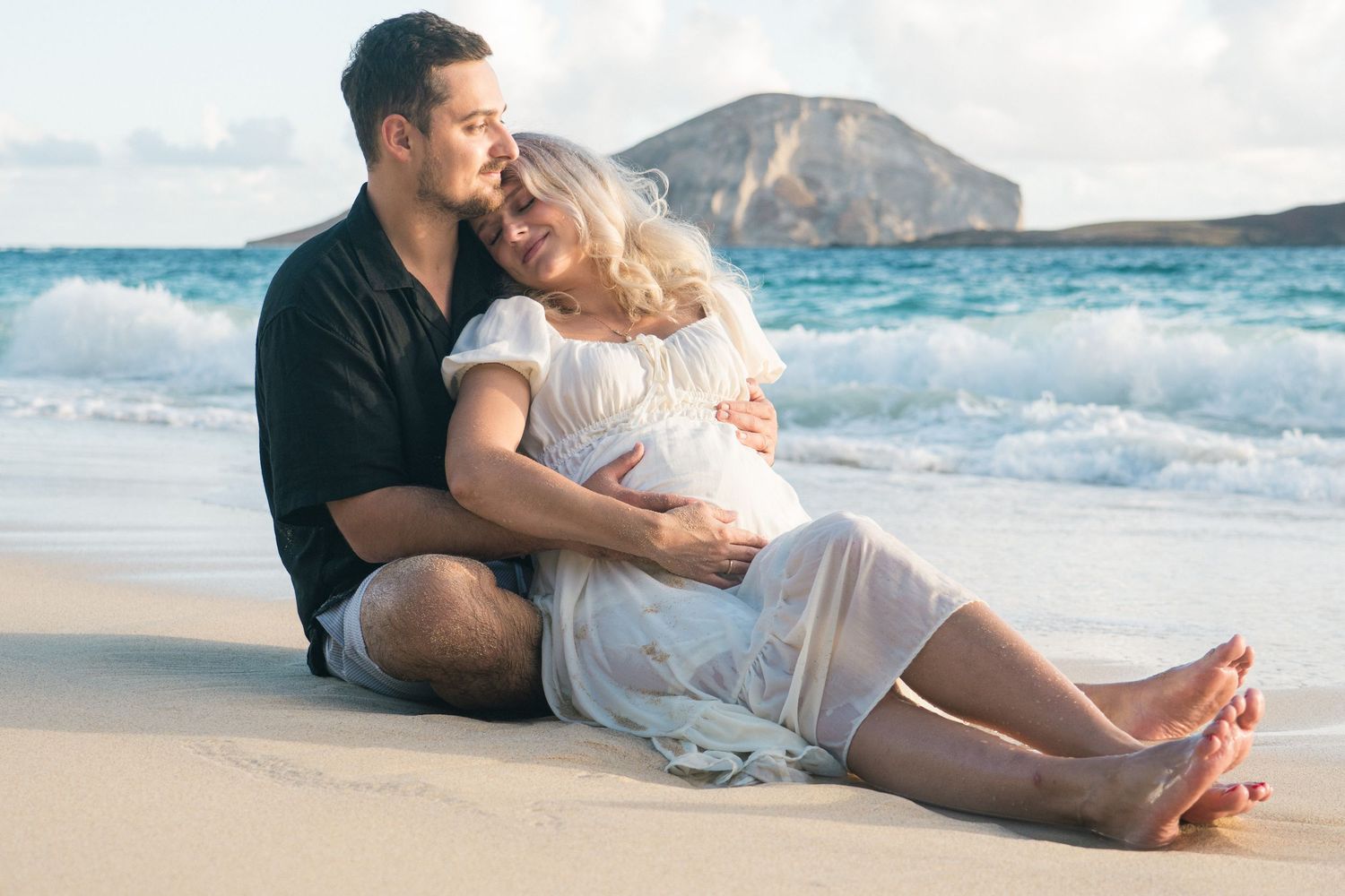 Couple sitting on sandy beach, pregnant woman in white dress embraced by man in black shirt, ocean waves and island backdrop.