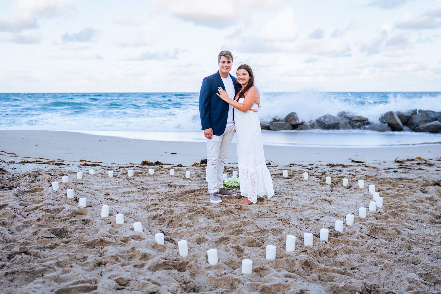 Newlywed couple posing inside a heart-shaped candle arrangement on a sandy beach with ocean waves behind them.