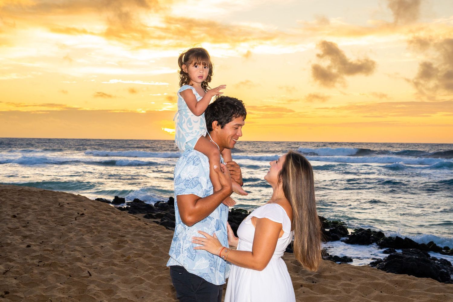 Family of three enjoying a beautiful sunset on a Hawaiian beach, with a young girl sitting on her father's shoulders.
